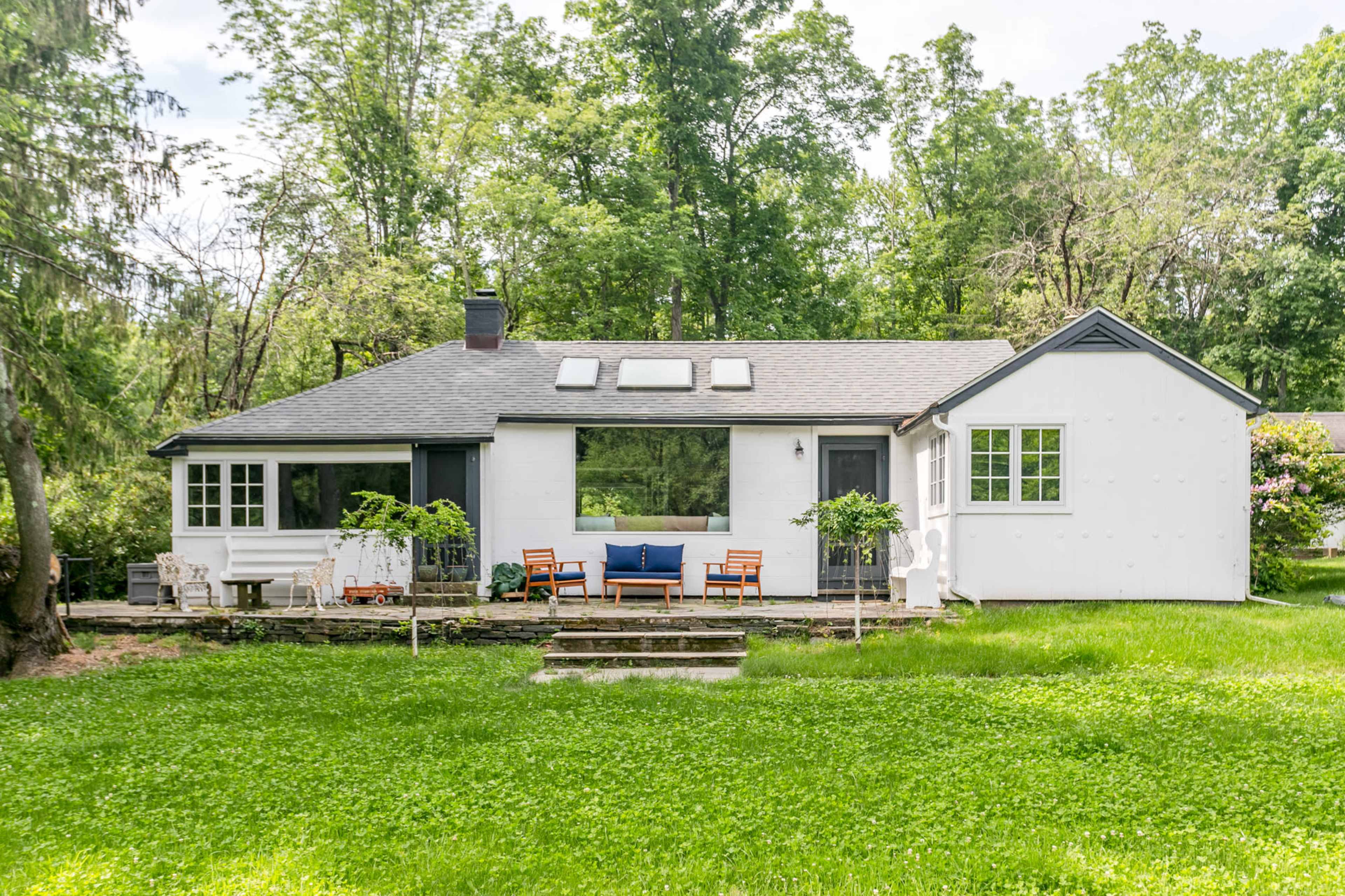 A single-story house with a combination of white and dark gray exterior is surrounded by greenery, featuring a front porch with seating and large windows.