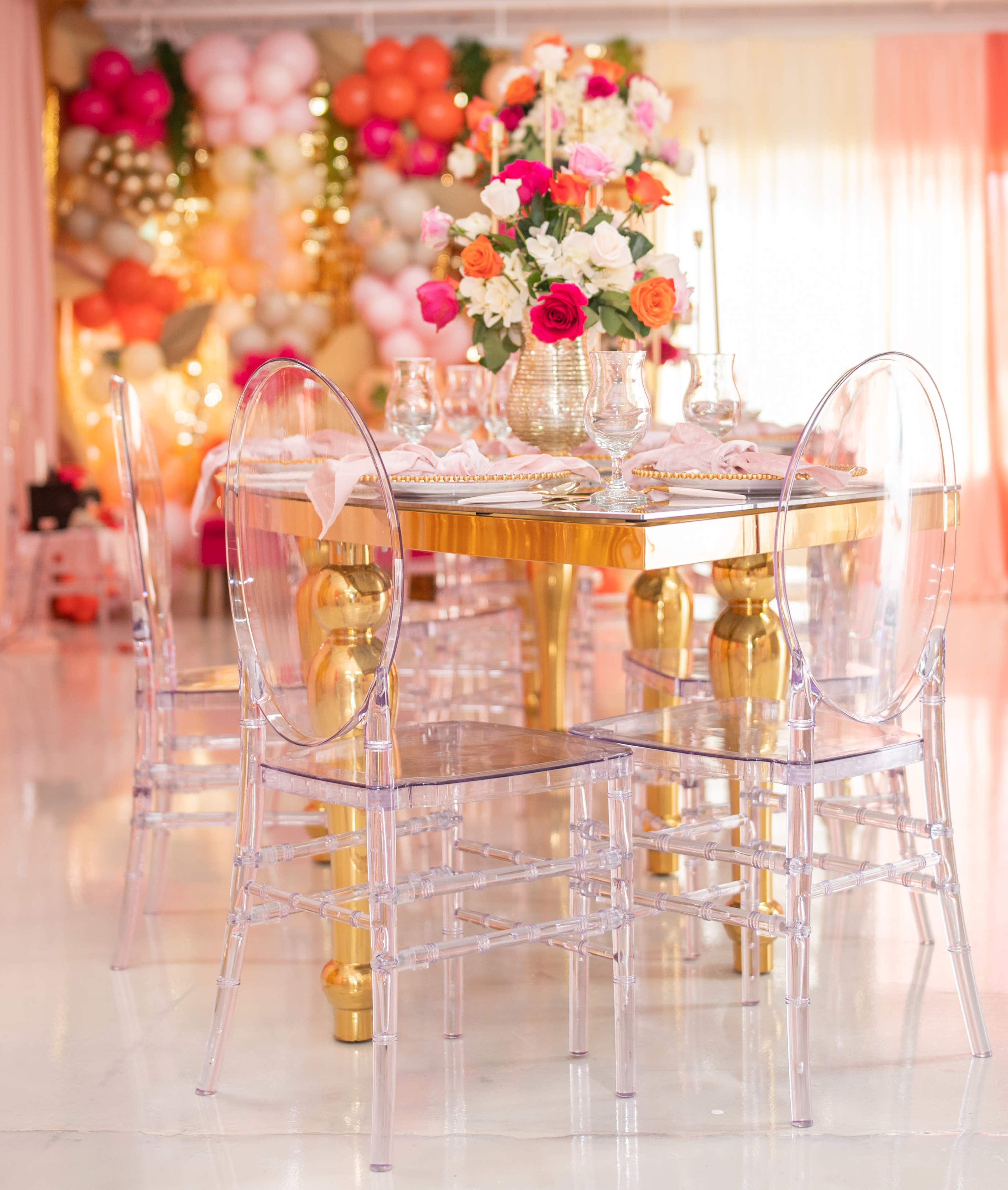 A glass table with a floral centerpiece is set with transparent chairs in a brightly lit room adorned with decorative balloons.
