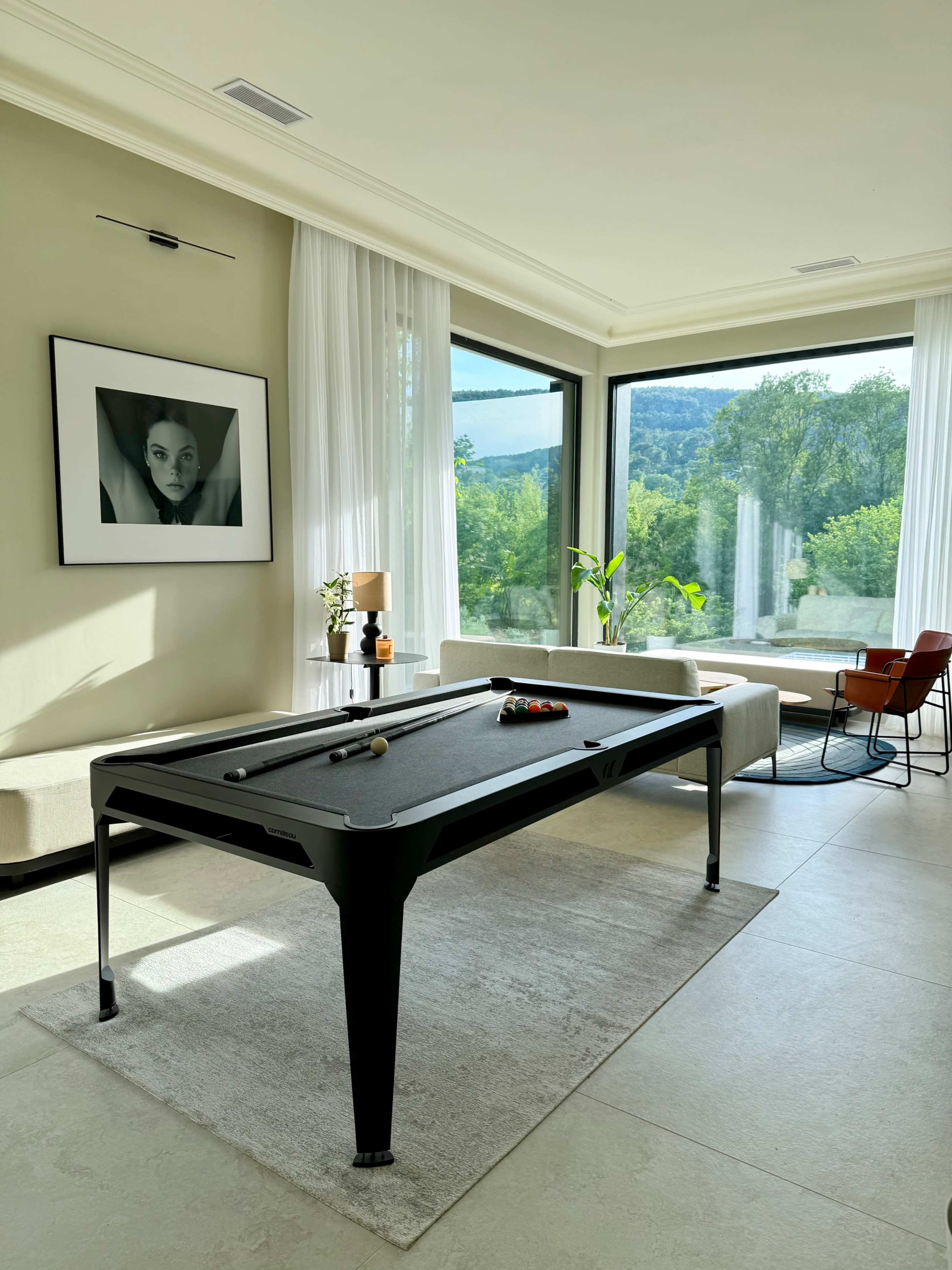 A modern interior features a billiards table in front of large windows with scenic views, complemented by a sofa and an armchair.