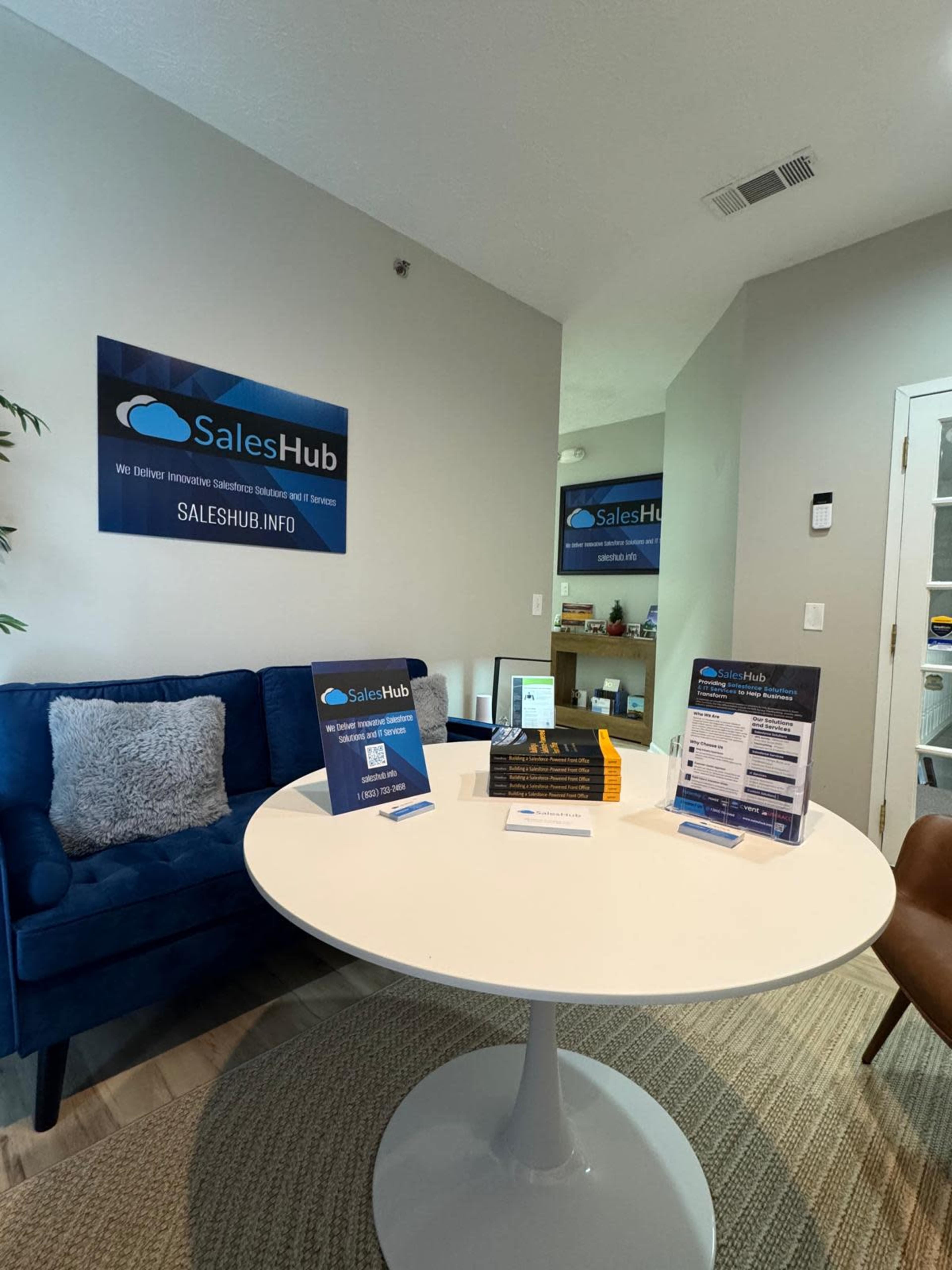 The image shows a modern office space featuring a blue couch, a round white table with promotional materials, and walls adorned with a sign for SalesHub.