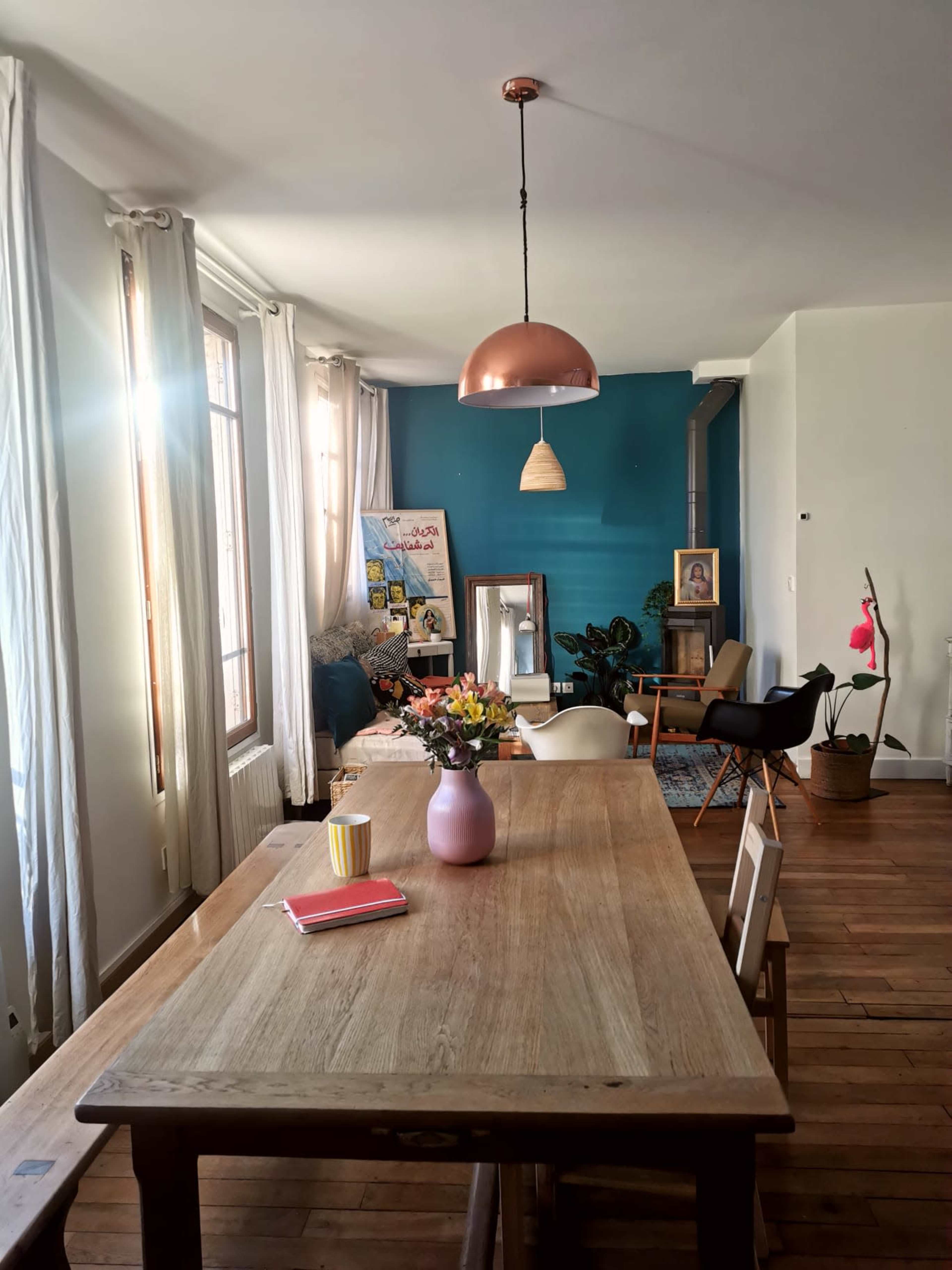 A dining area features a wooden table with a pink vase of flowers, booklets, and a yellow cup under a copper pendant light, with a blue accent wall and chairs in the background.