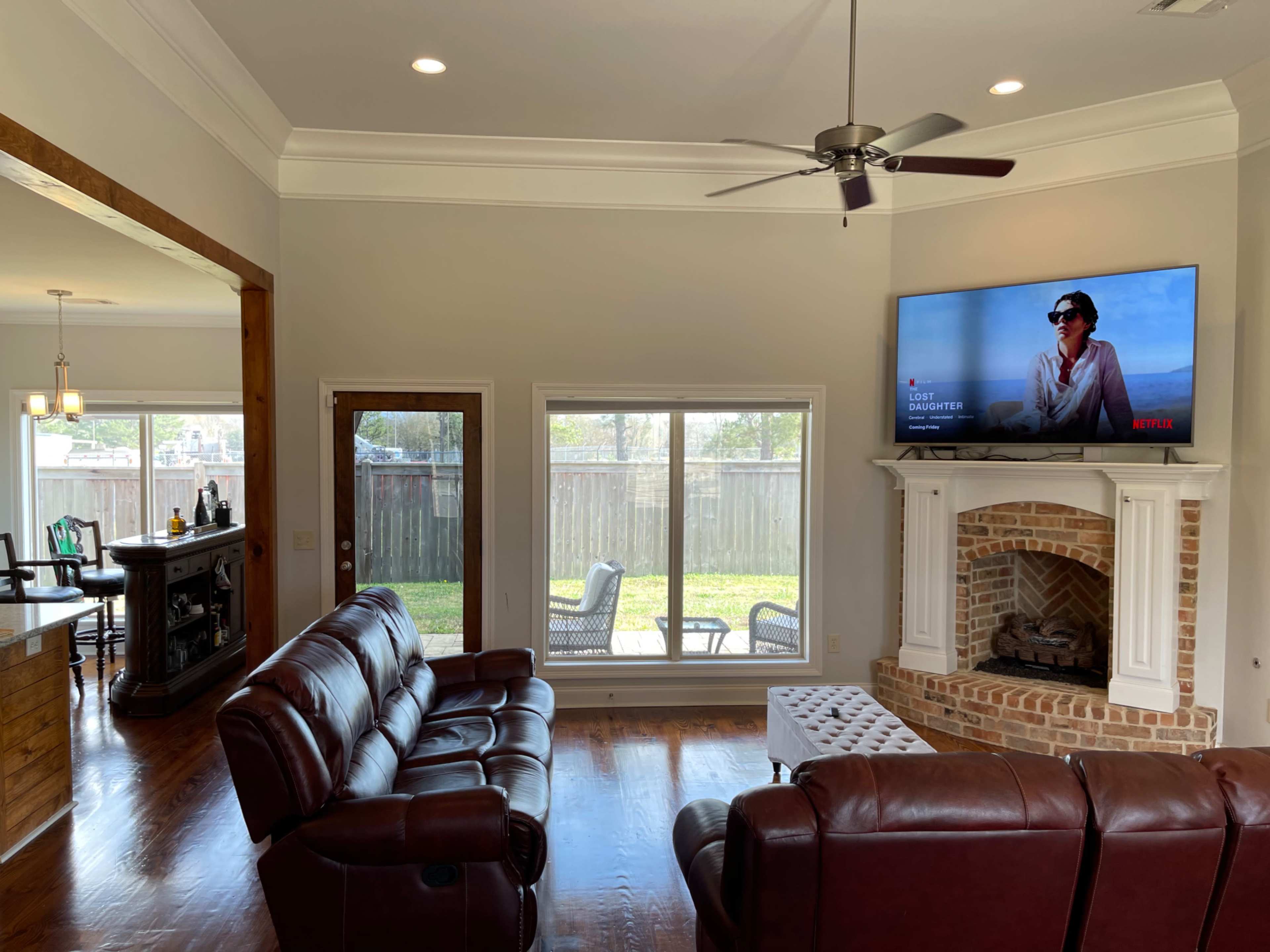 The living room features brown leather sofas, a brick fireplace, a television mounted above the mantel, and large windows overlooking a backyard.