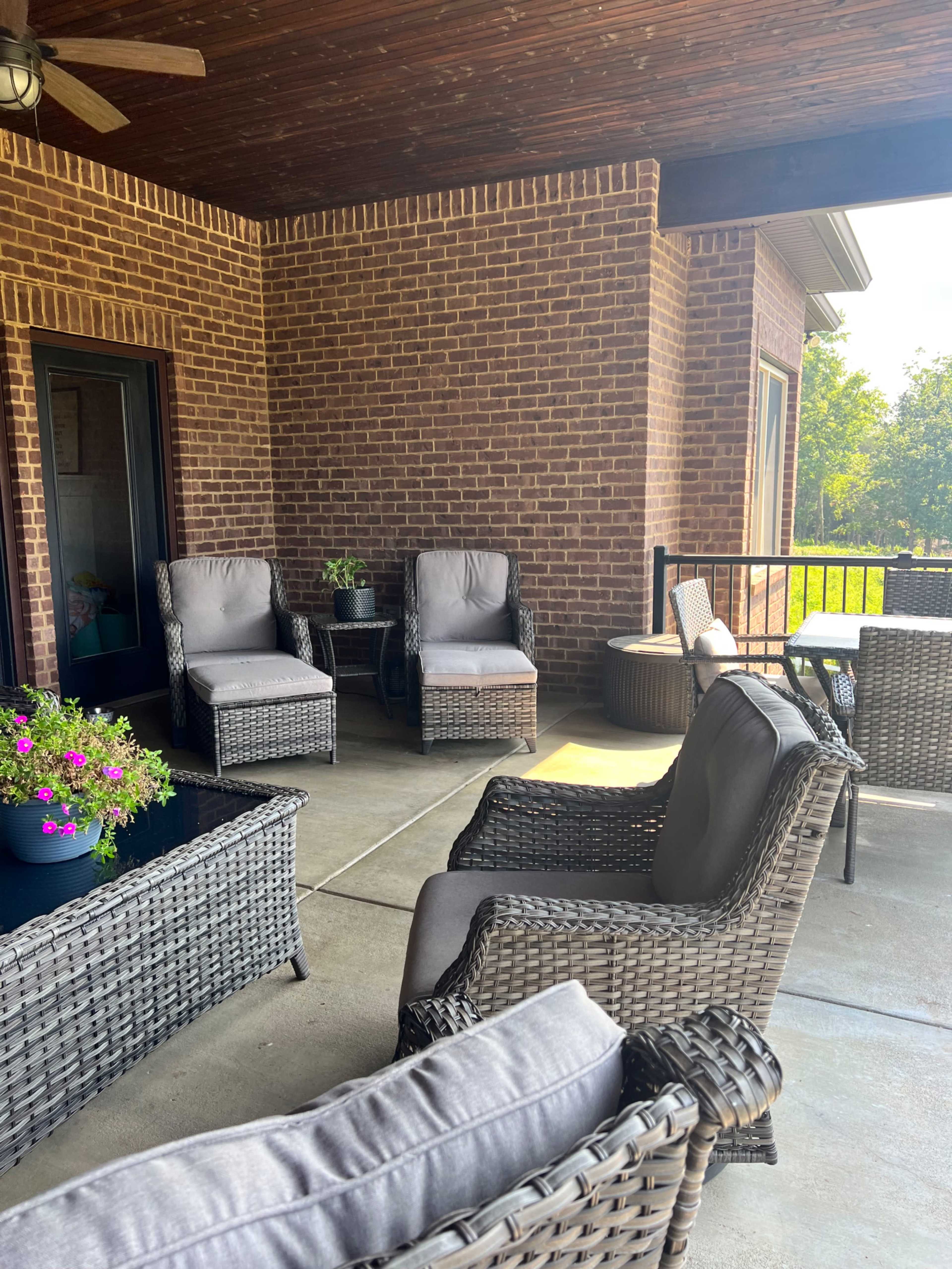The image shows a patio area with wicker furniture, including chairs and a coffee table, surrounded by brick walls and sunlight filtering in.