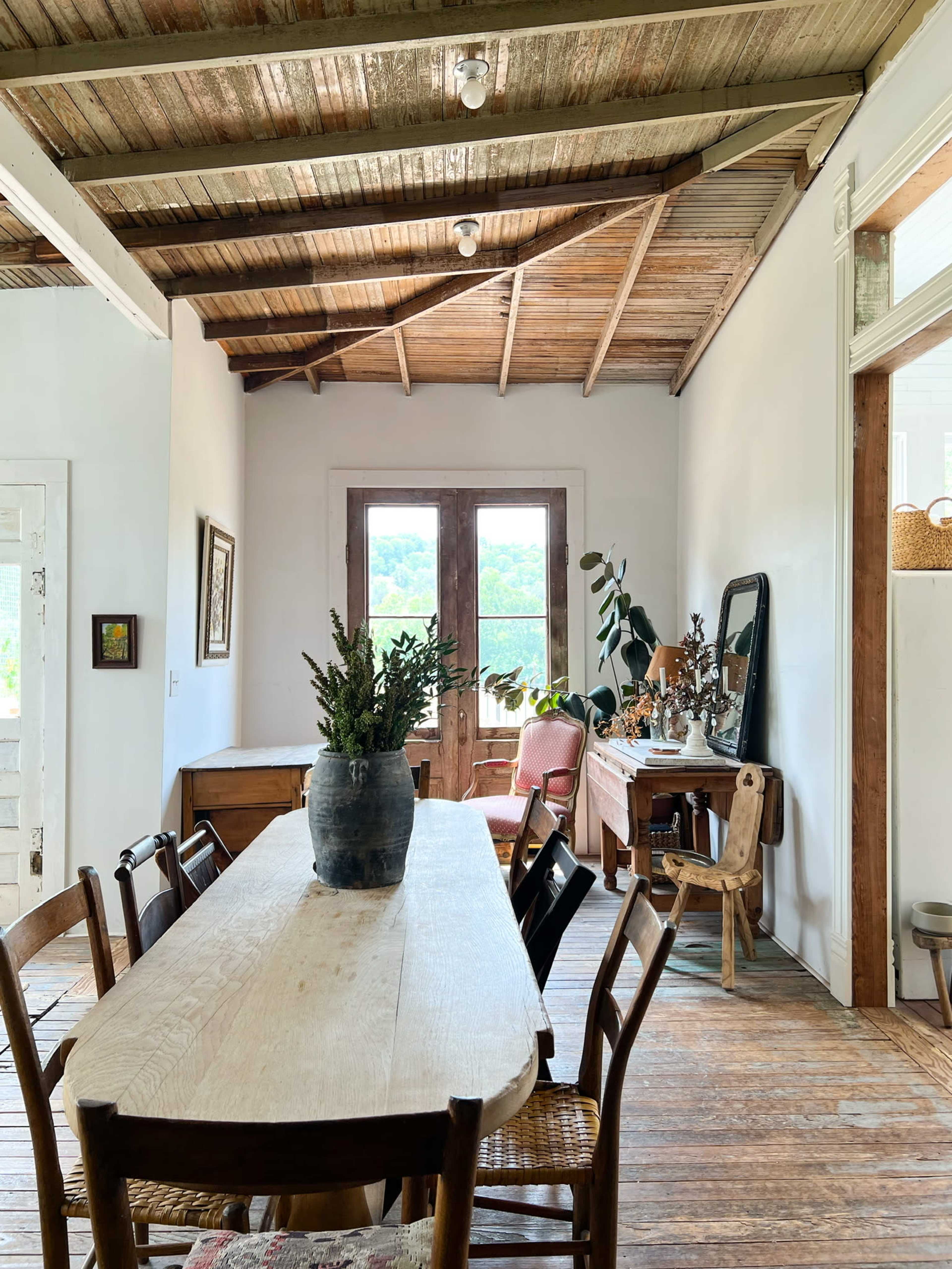 A long wooden dining table is centered in a room with a whitewashed wall, featuring a door that leads to a view of greenery outside.