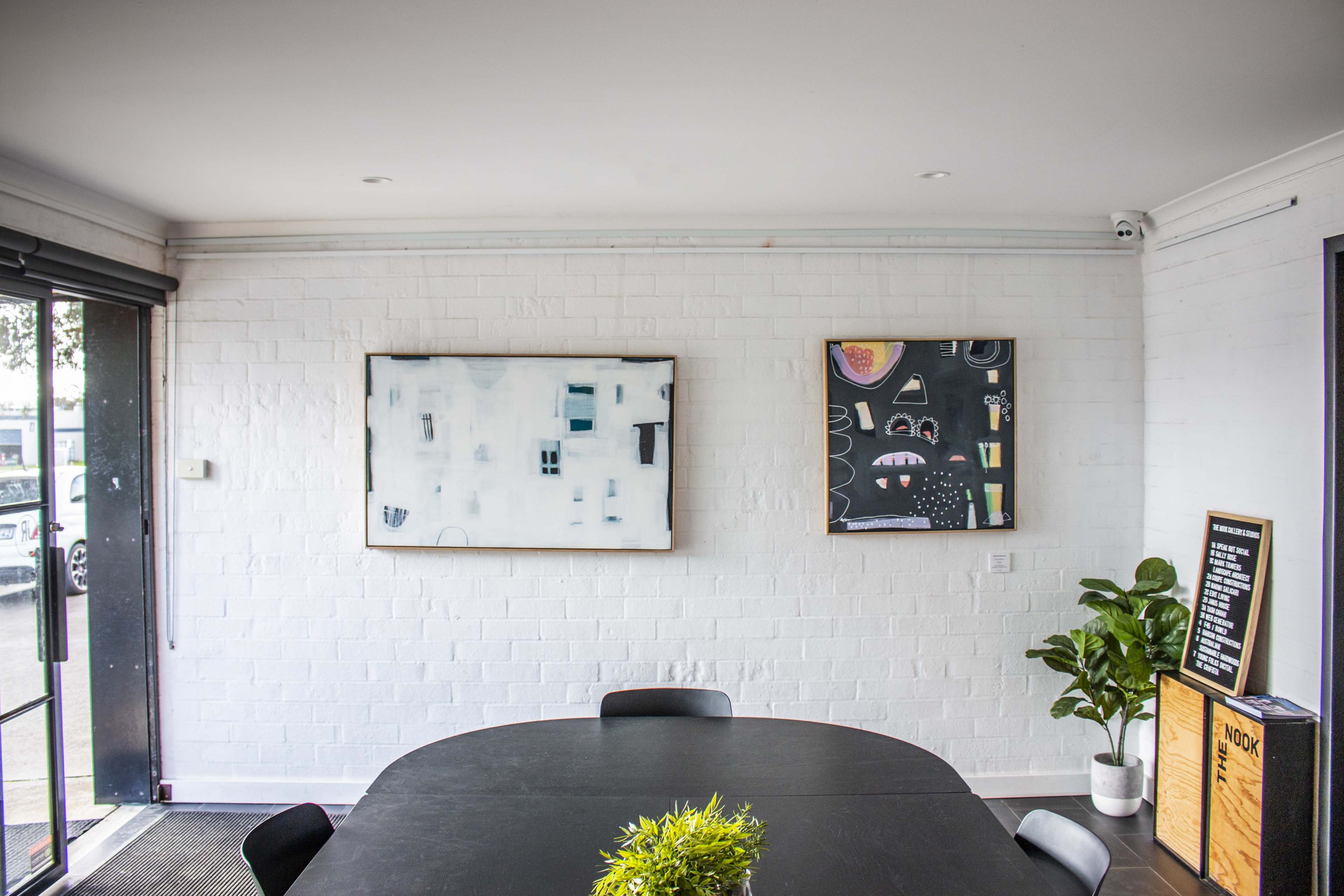 A modern interior featuring a round black table, two abstract paintings on the white brick wall, and a potted plant in the corner.