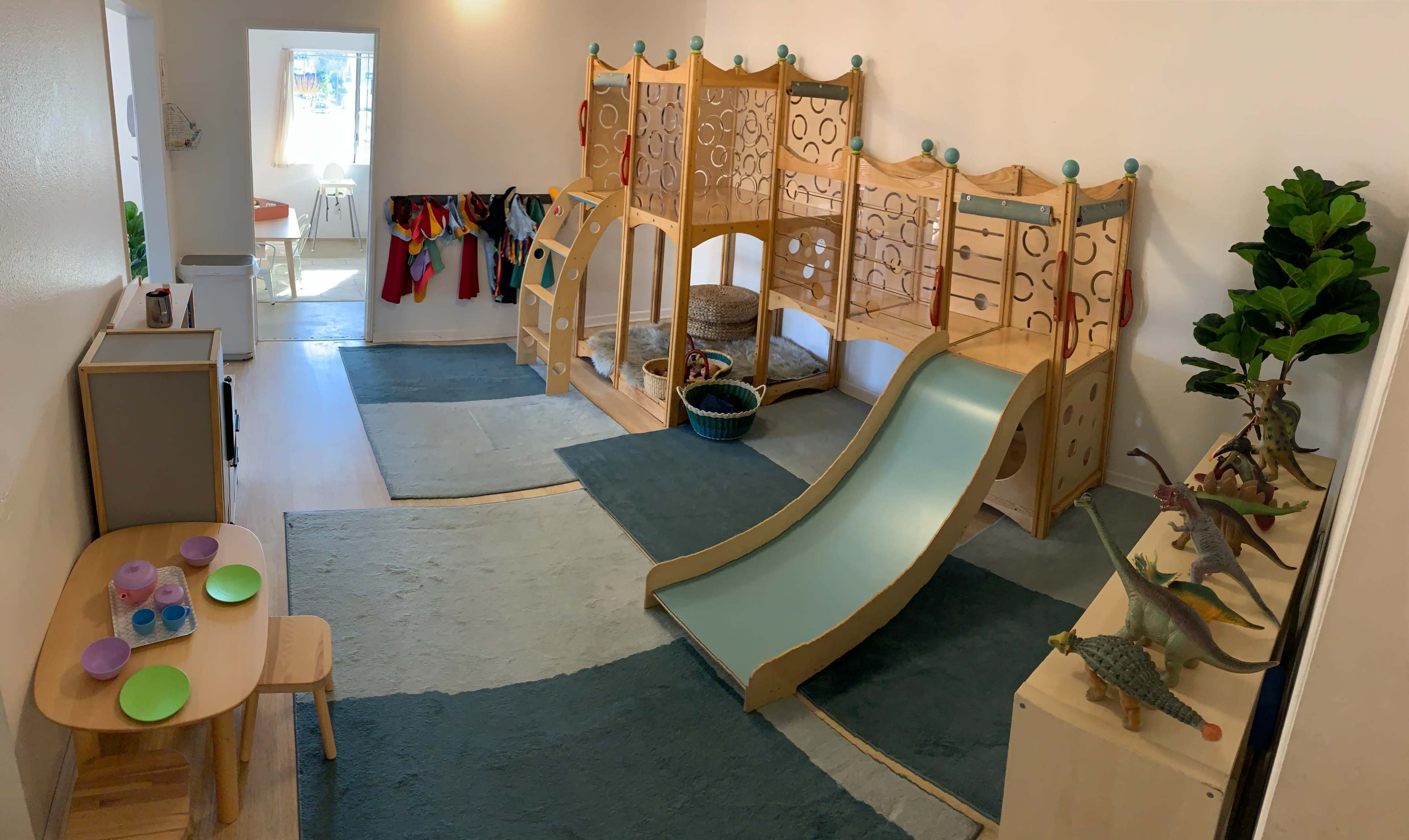 A brightly lit playroom featuring a wooden climber with a slide, soft blue rugs, and a small table with plates in one corner.