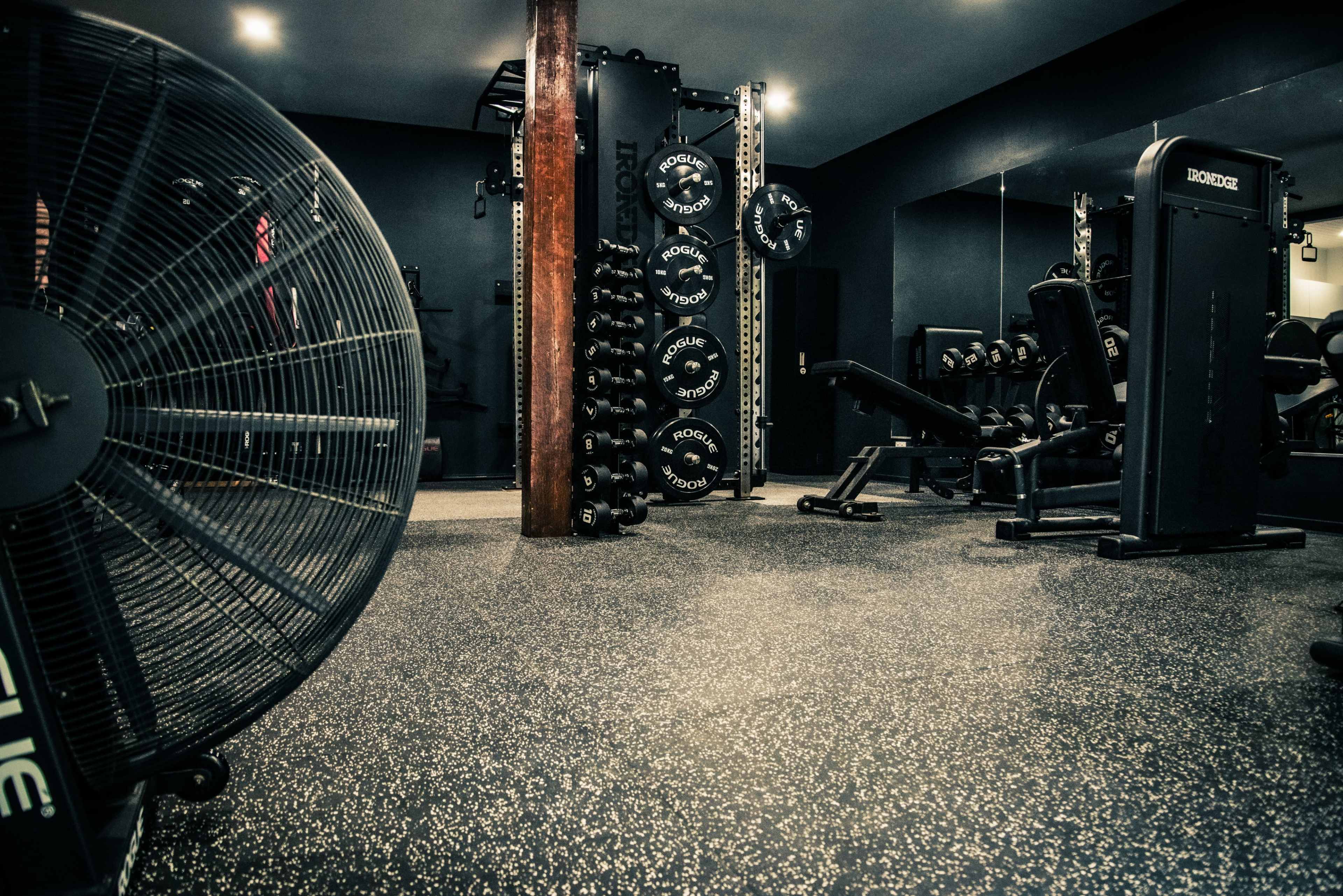 The image shows a modern gym interior featuring workout equipment, free weights, and a large floor fan.