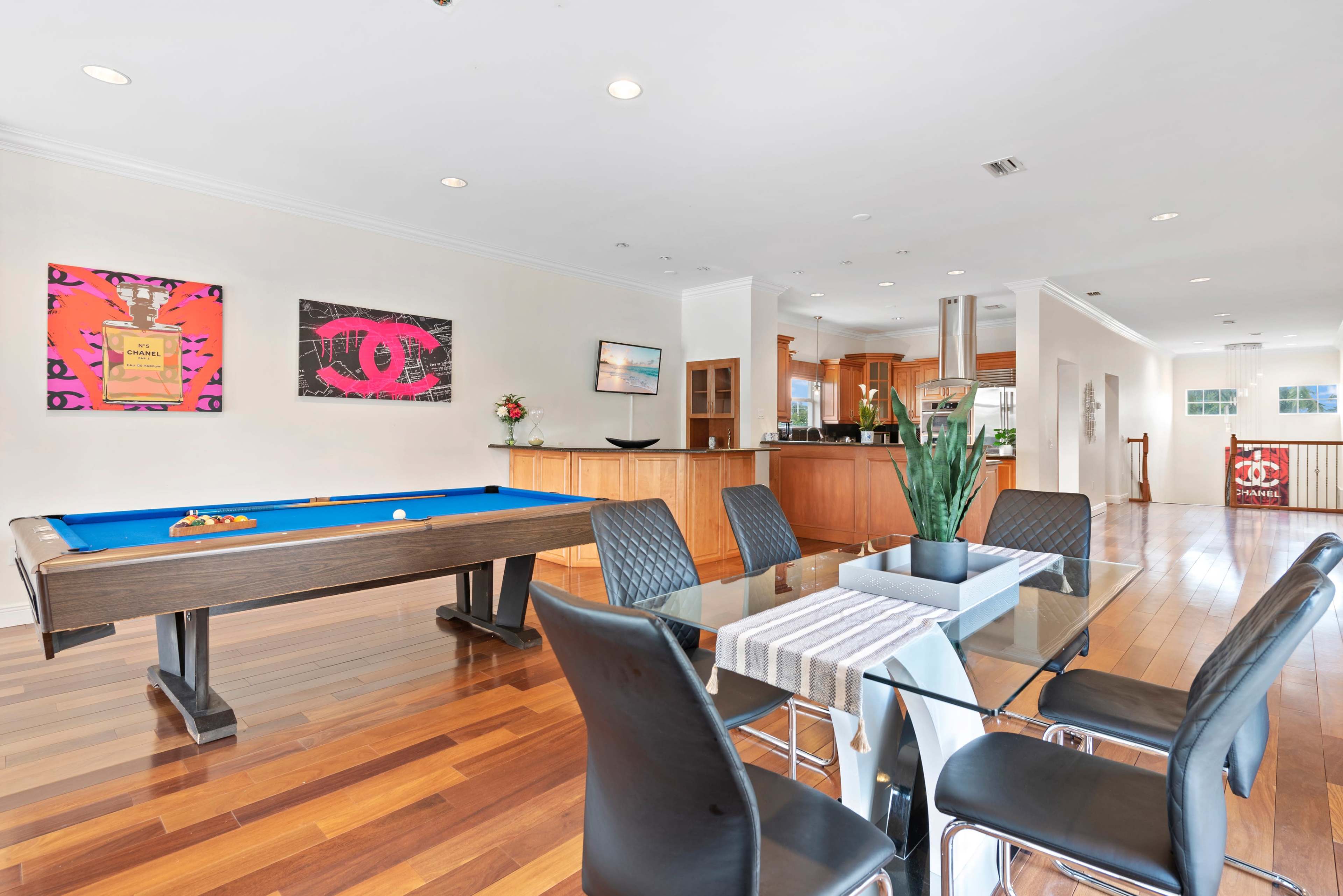 A modern open-concept living space featuring a pool table, dining area with a glass table, and a kitchen in the background.