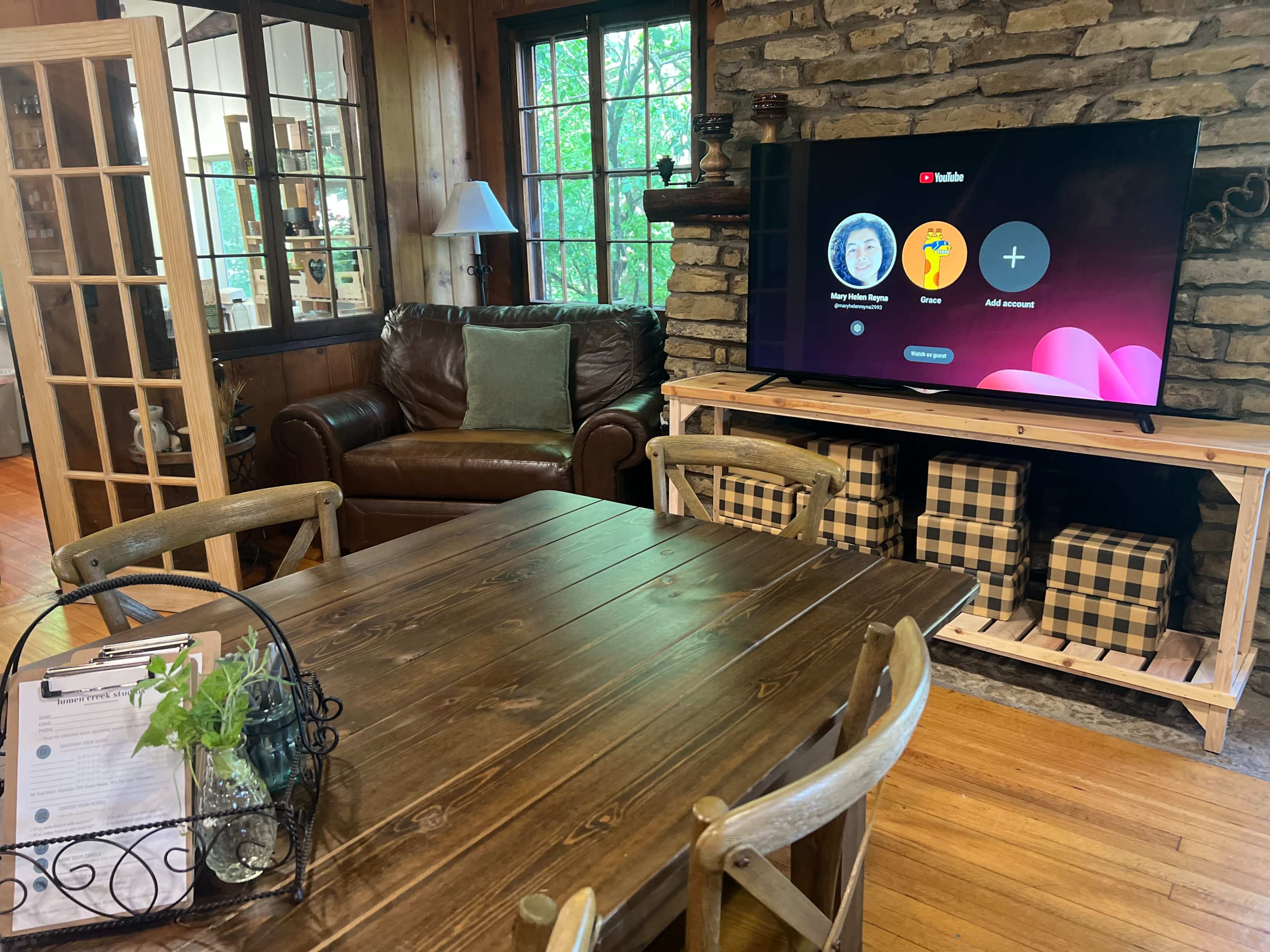 A cozy living room features a wooden table, a leather couch, and a television displaying a YouTube interface against a stone wall.