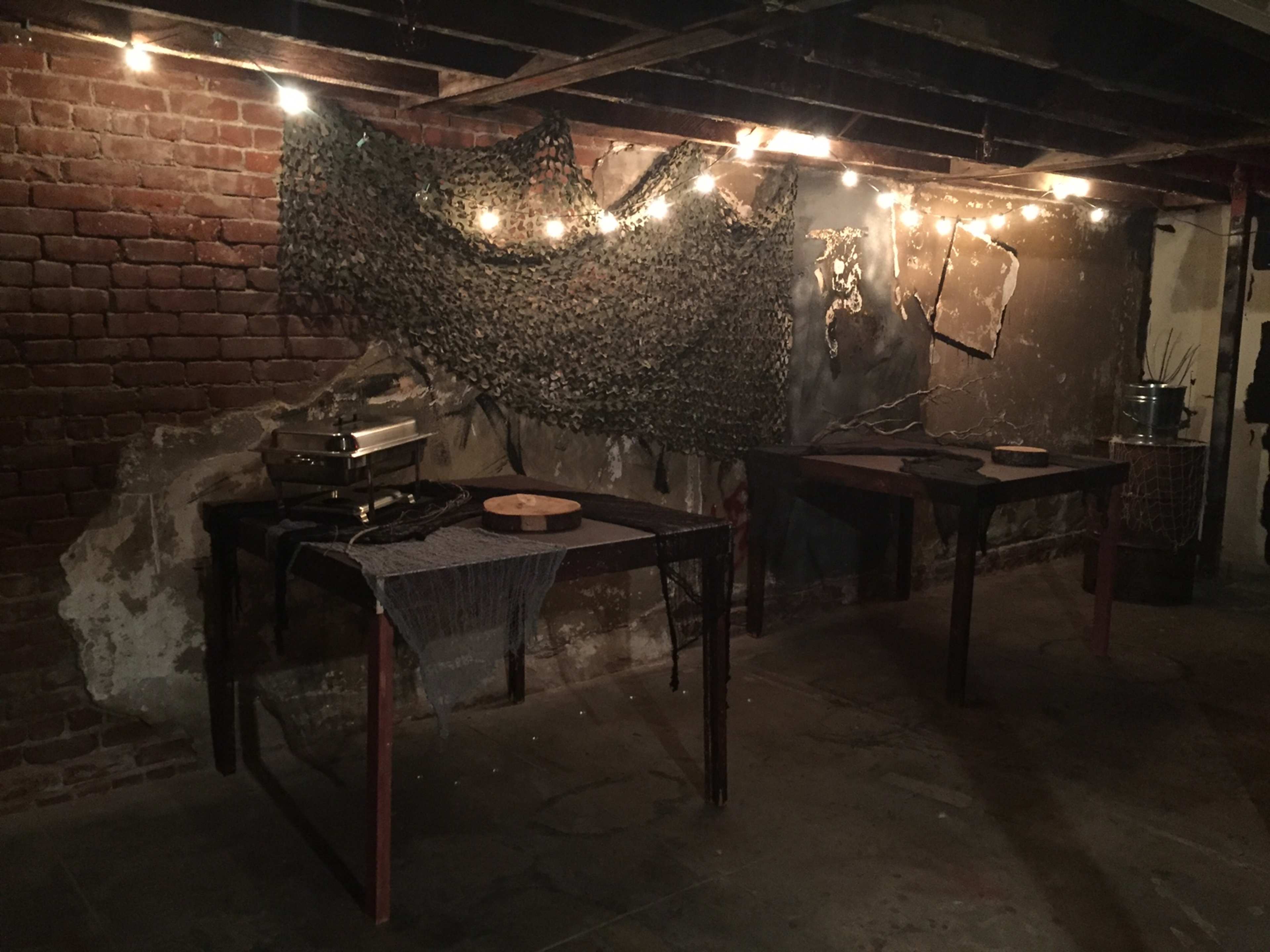 A dimly lit basement with two wooden tables, a backdrop of camouflage netting, and string lights overhead illuminating the brick and concrete walls.