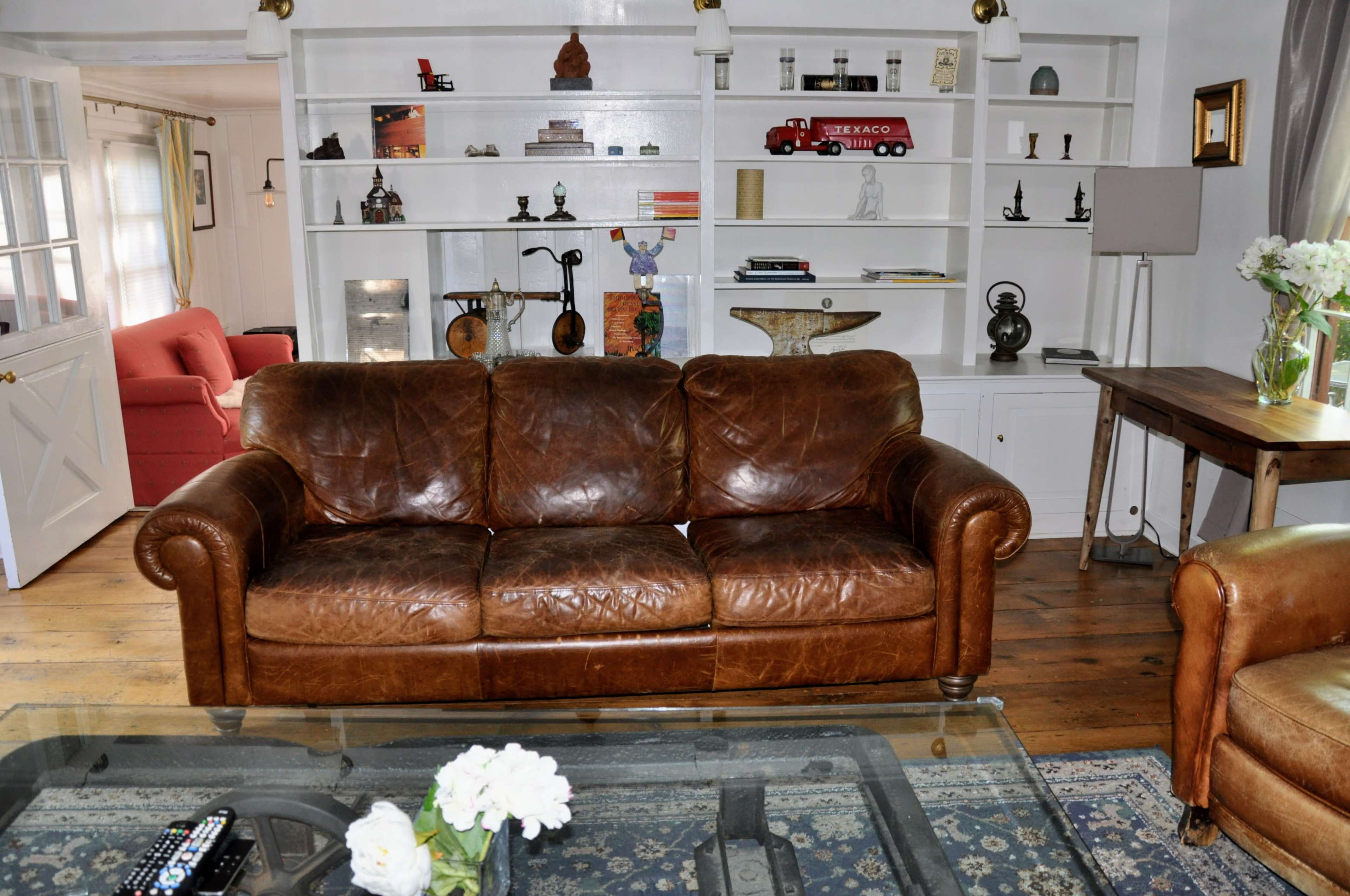 The image shows a brown leather sofa in a living room with white shelves displaying various decorative items and a glass coffee table with flowers.