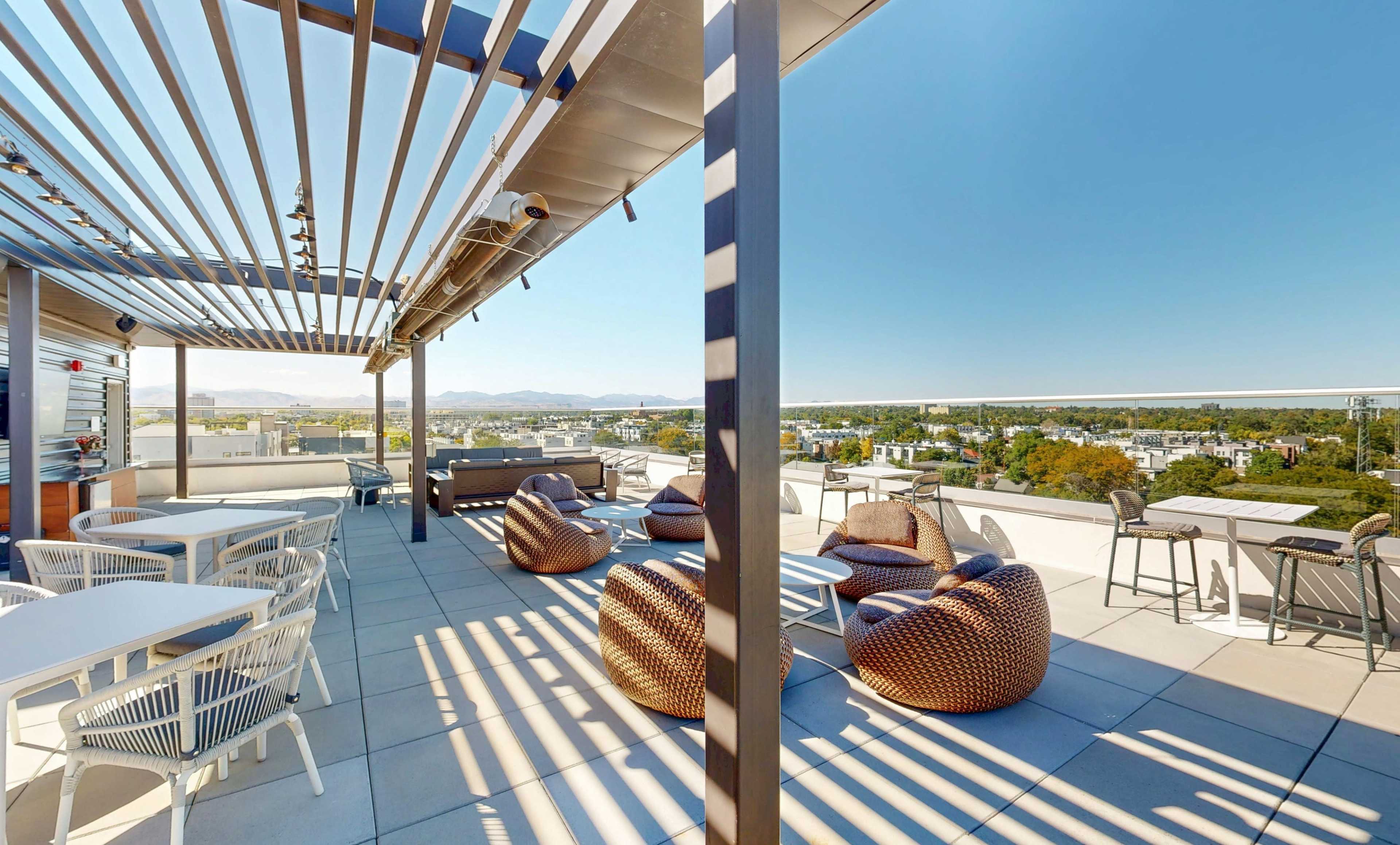 The image shows a rooftop terrace with seating arrangements, including lounge chairs and tables, overlooking a landscape of buildings and hills under a clear blue sky.