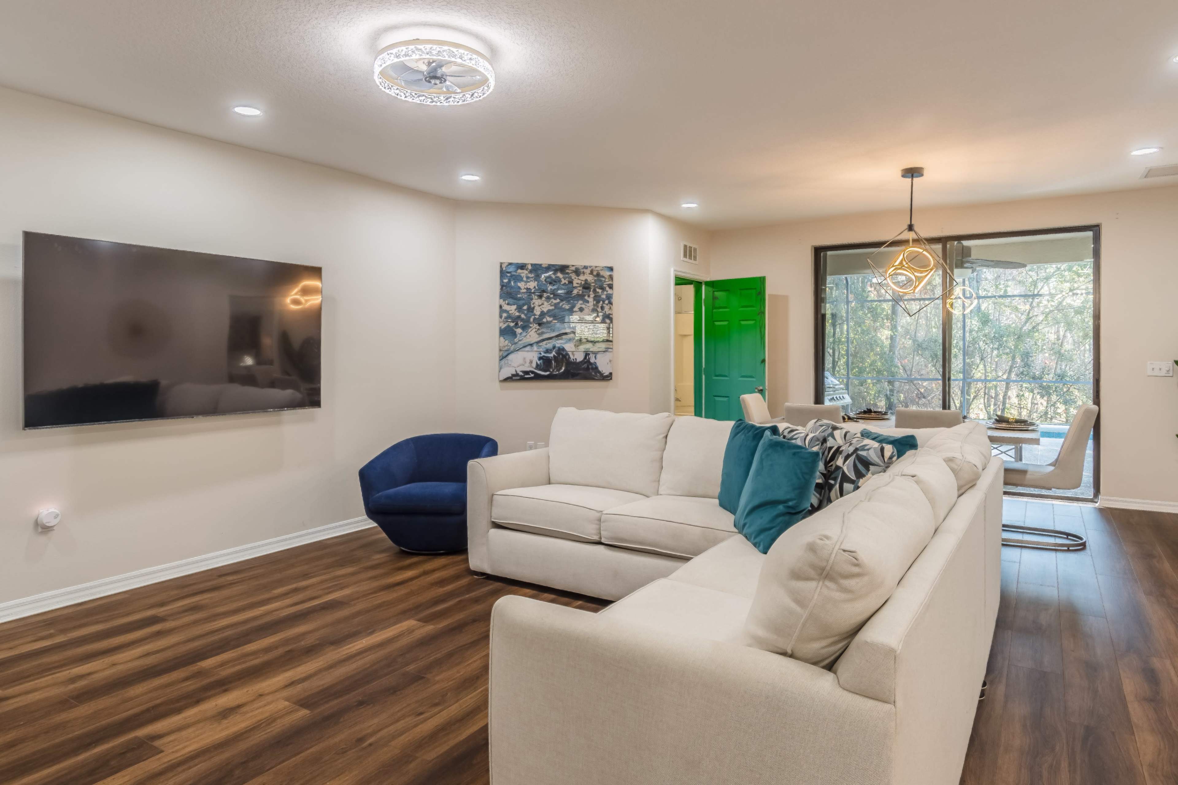 A modern living room features a light-colored sectional sofa, a dark blue accent chair, and a wall-mounted television, with large windows providing views of greenery outside.