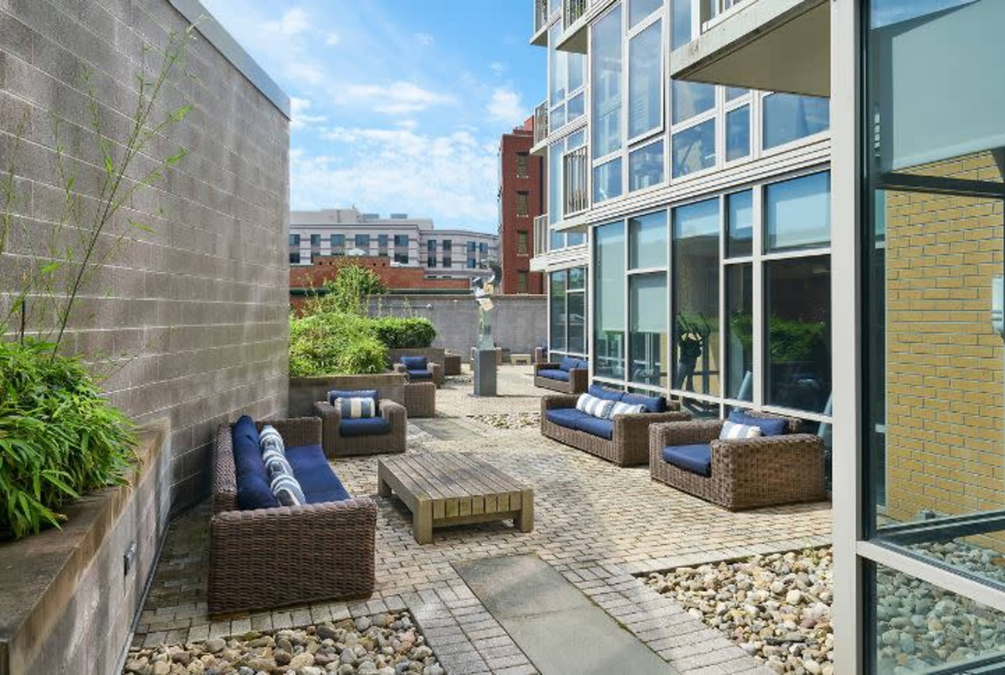 The image shows an outdoor lounge area with wicker furniture, including sofas and a coffee table, surrounded by greenery and buildings in the background.