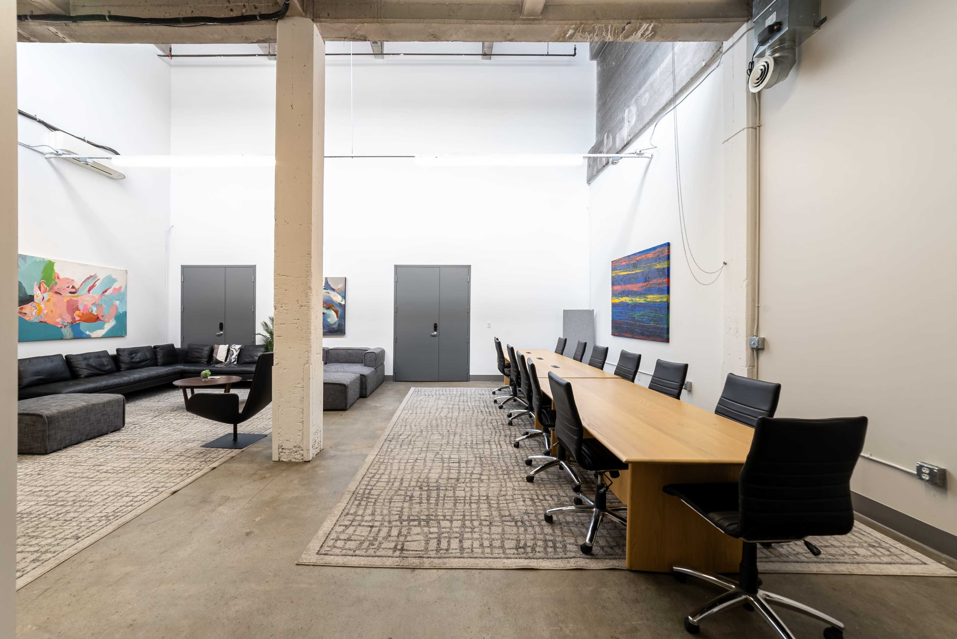 A spacious office interior featuring a long wooden conference table surrounded by black chairs, with a lounge area containing a dark couch and colorful artworks on the walls.