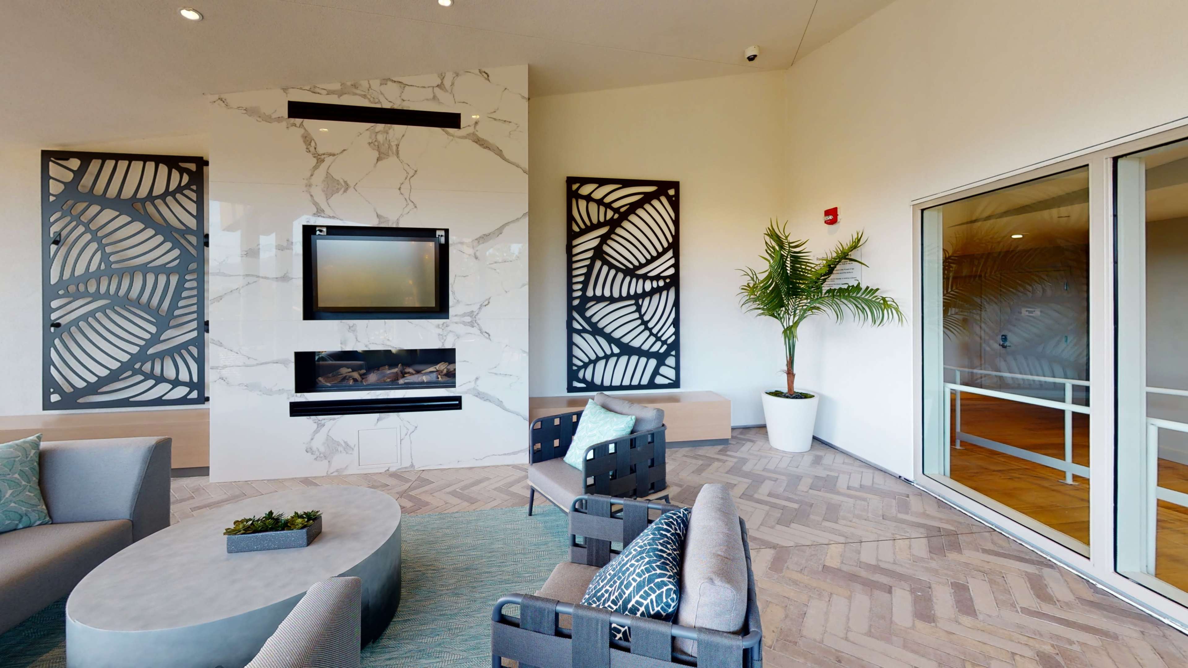 The image shows a modern lounge area featuring a marble fireplace, patterned wall art, and comfortable seating arranged around a circular table.