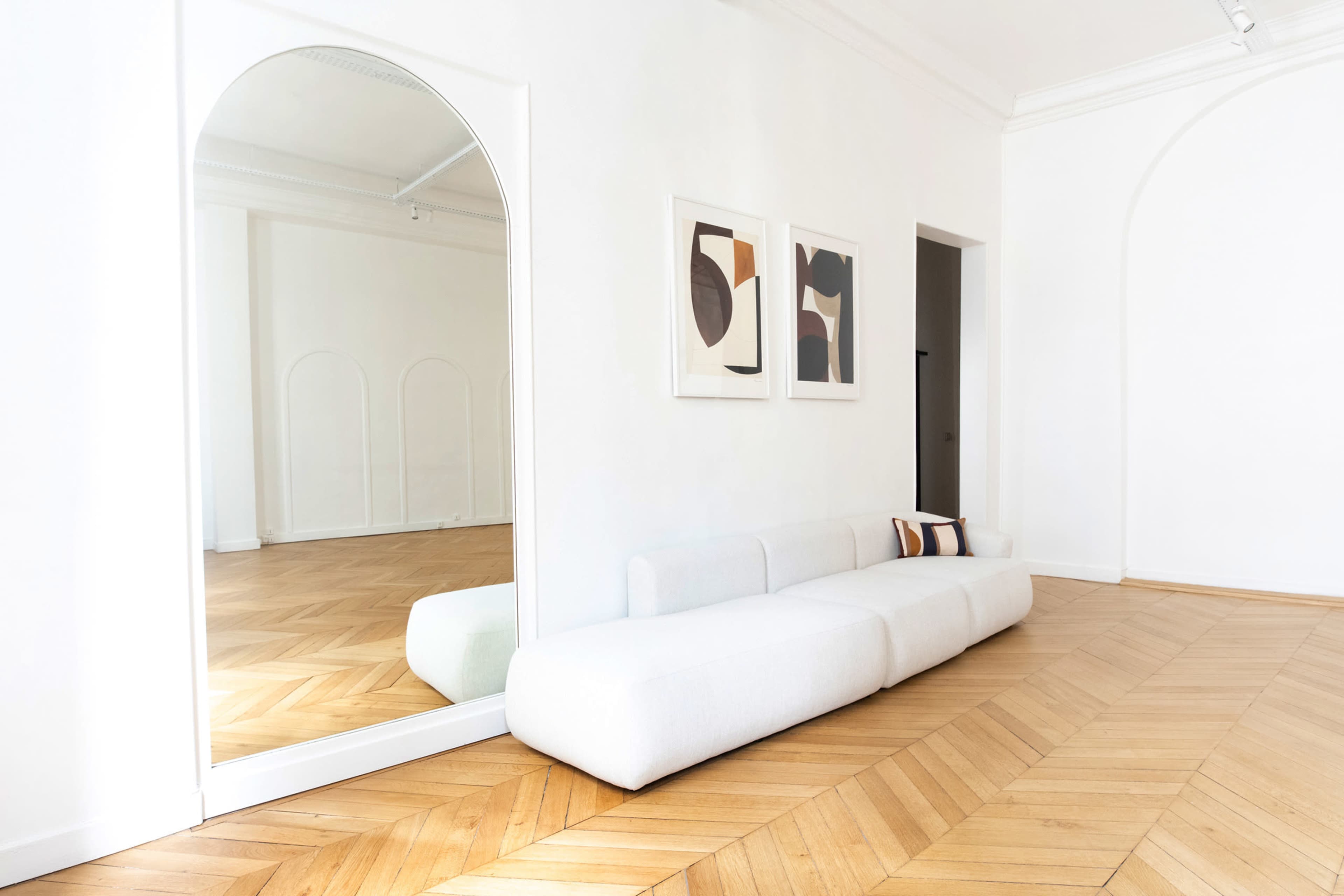 A minimalist room with a light wooden floor, a white curved sofa, and a large arched mirror reflecting the space.