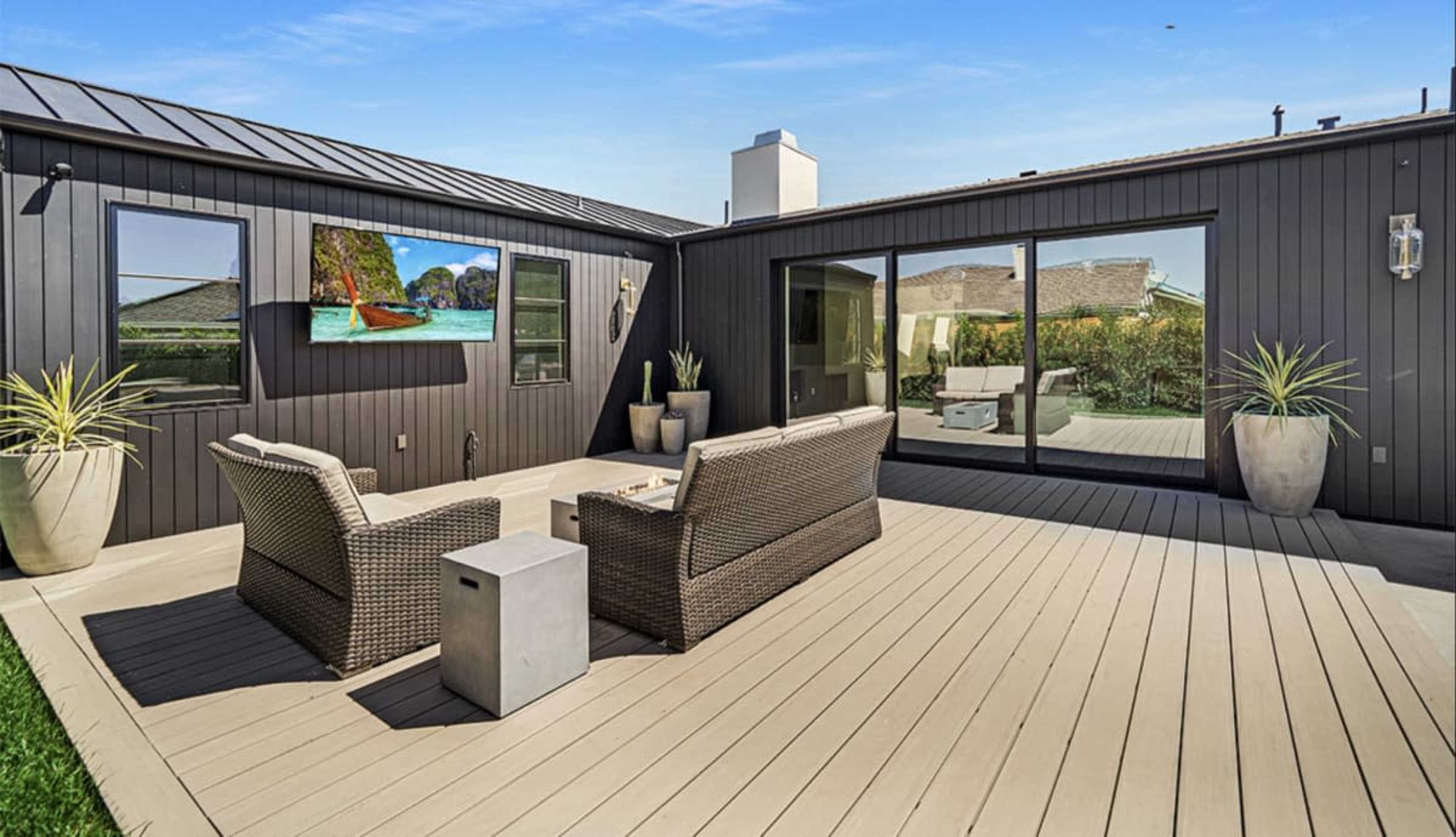 The image shows a modern outdoor patio with wicker furniture, a small table, and a wall-mounted TV, surrounded by large planters.