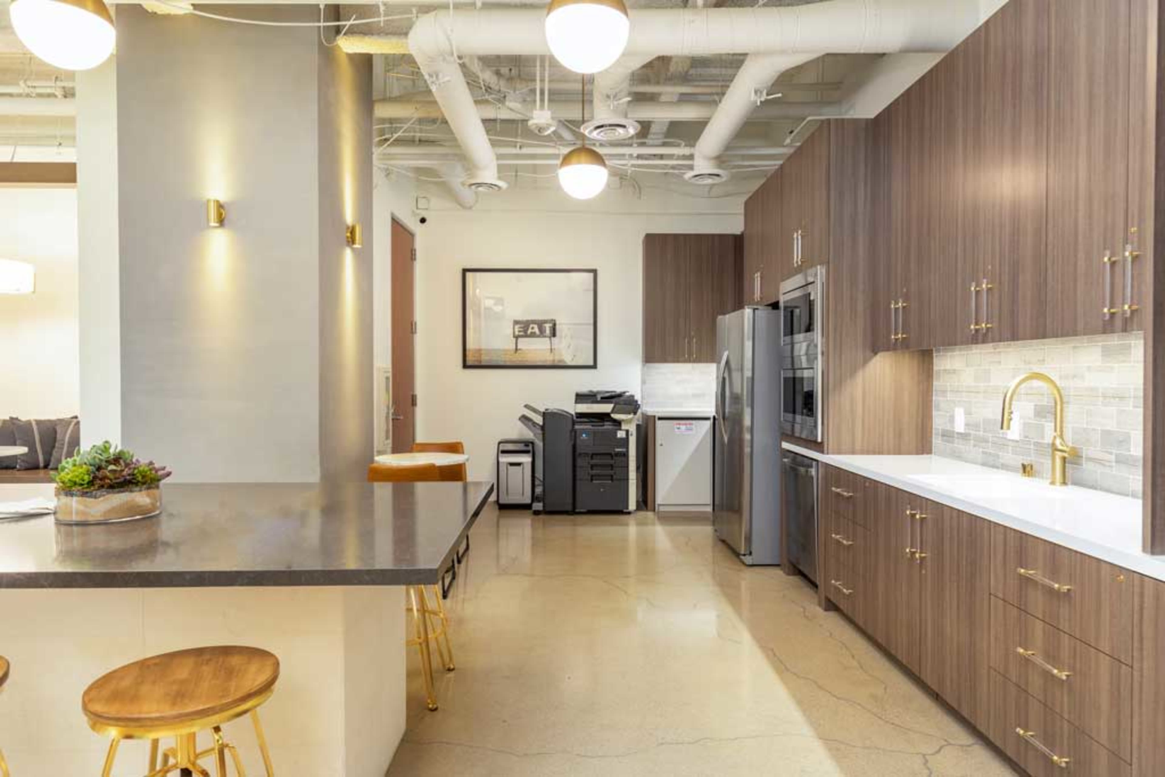 A modern kitchen with wooden cabinets, a large island with bar stools, and a small office space featuring a printer and a framed wall art.