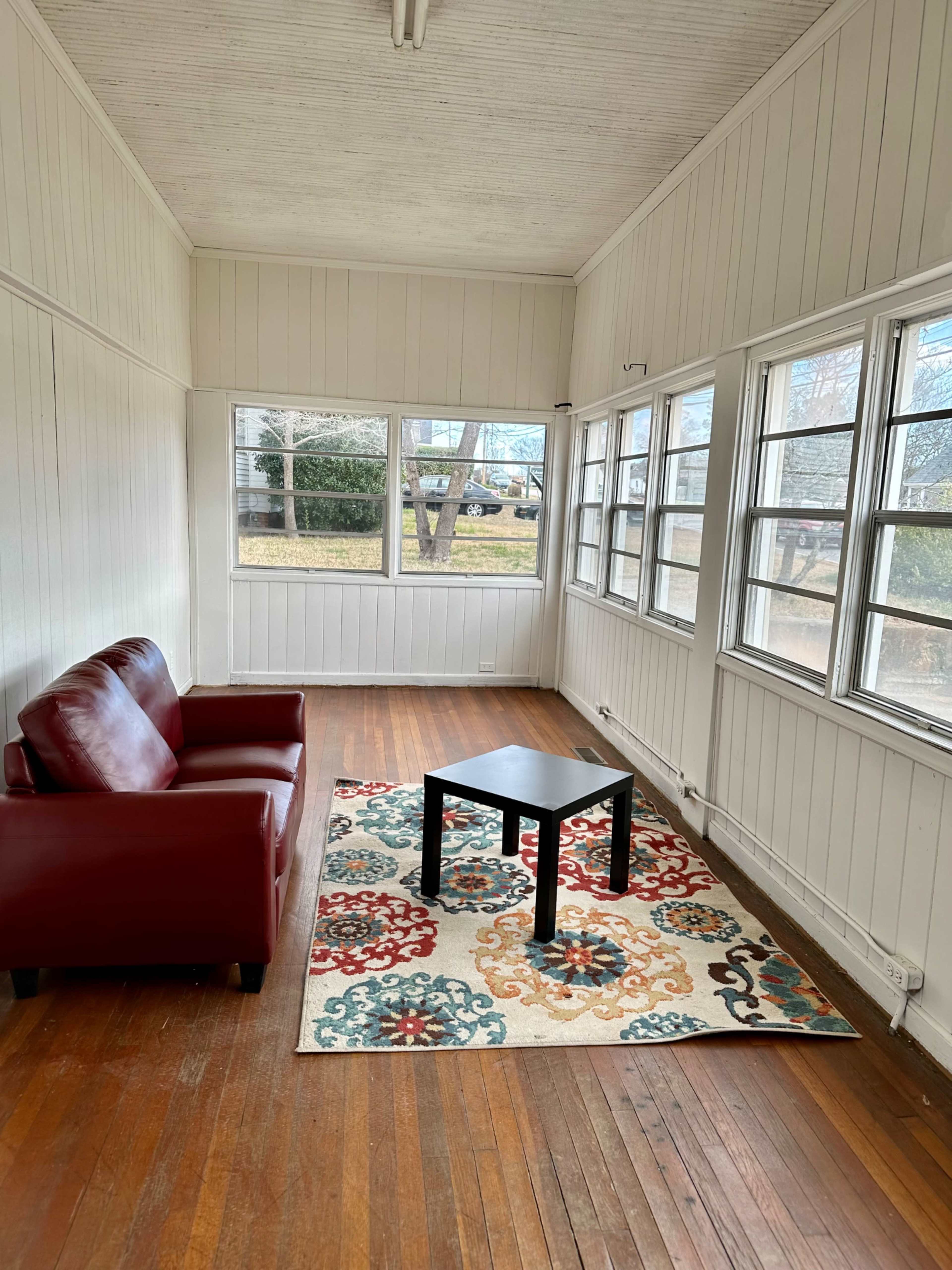 A small sunroom features a red sofa, a black coffee table, and a patterned area rug, surrounded by large windows.