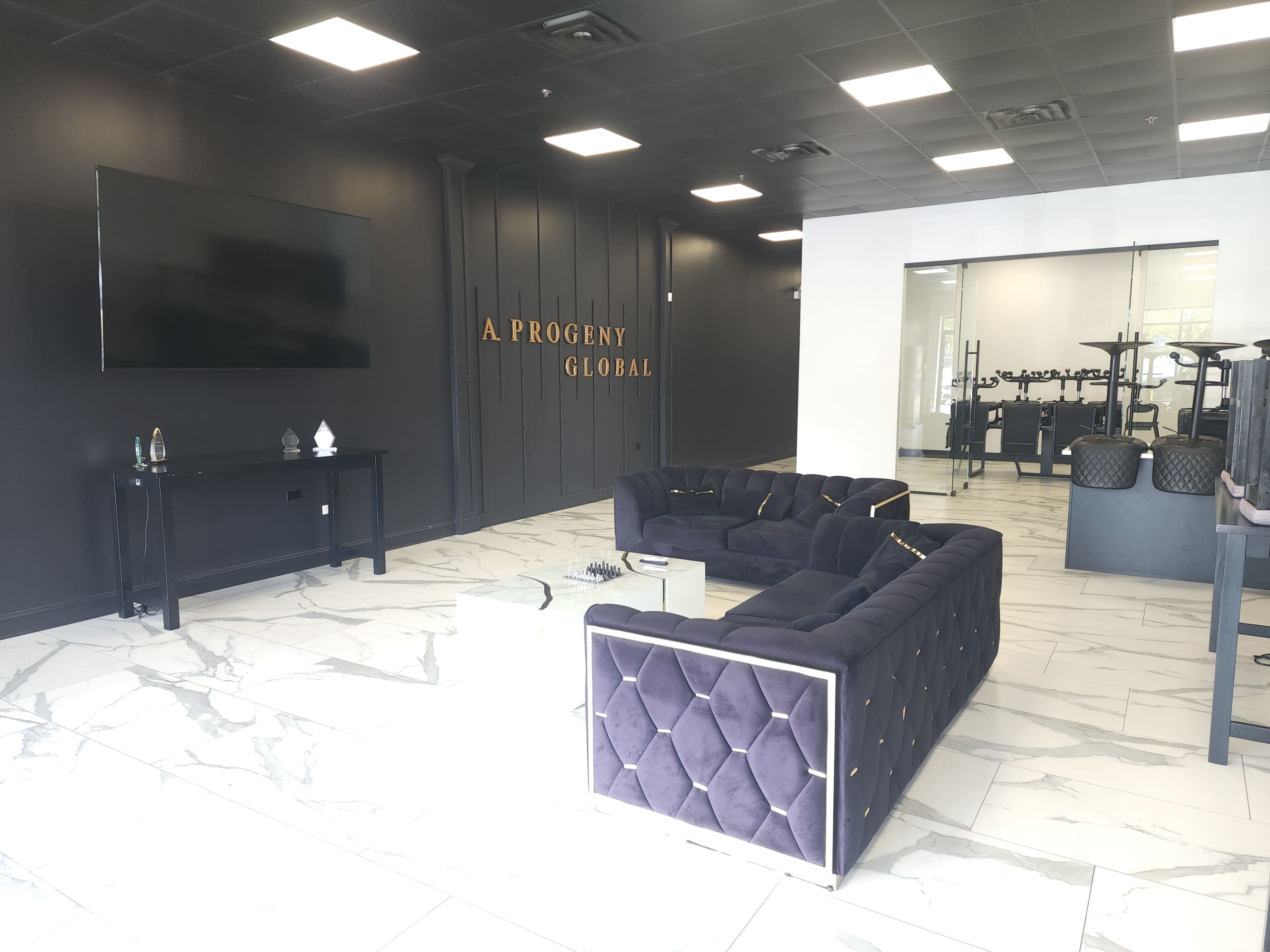 The image shows a modern reception area featuring a dark velvet sofa, a wall-mounted TV, and a sleek white space with a fitness area visible in the background.