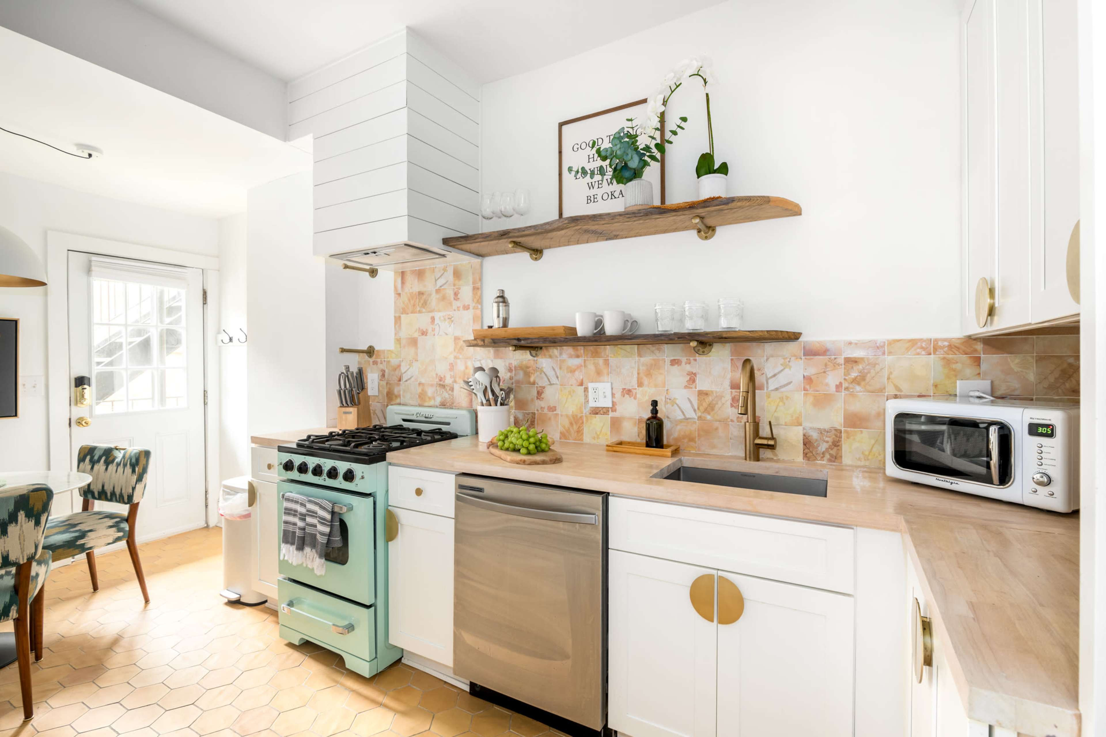 A modern kitchen features white cabinetry, an aqua stove, and open shelves displaying glassware and decor against a colorful tiled backsplash.