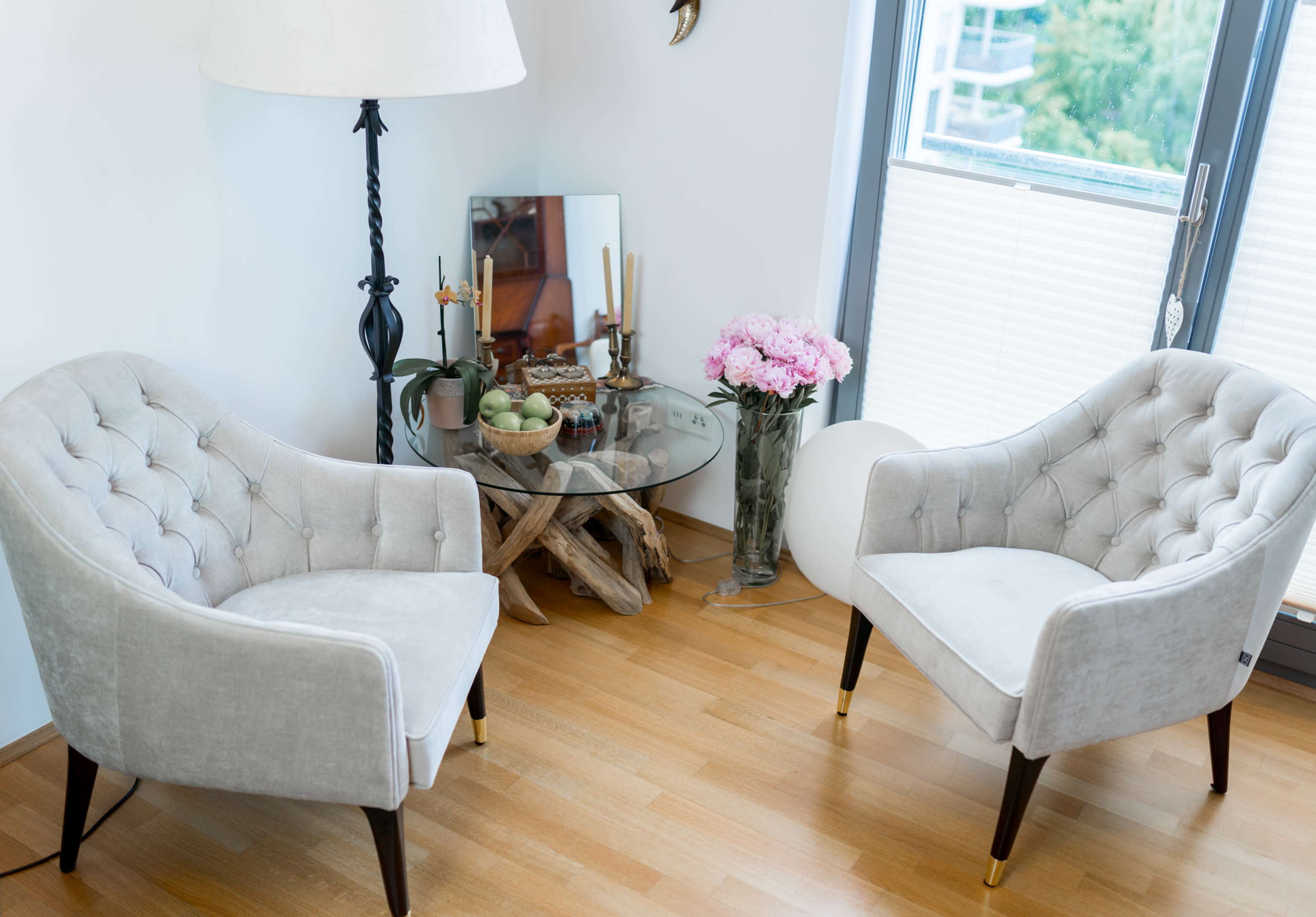 A cozy seating area with two tufted chairs, a glass table on a wooden stand, a small vase of pink flowers, and various decorative items.