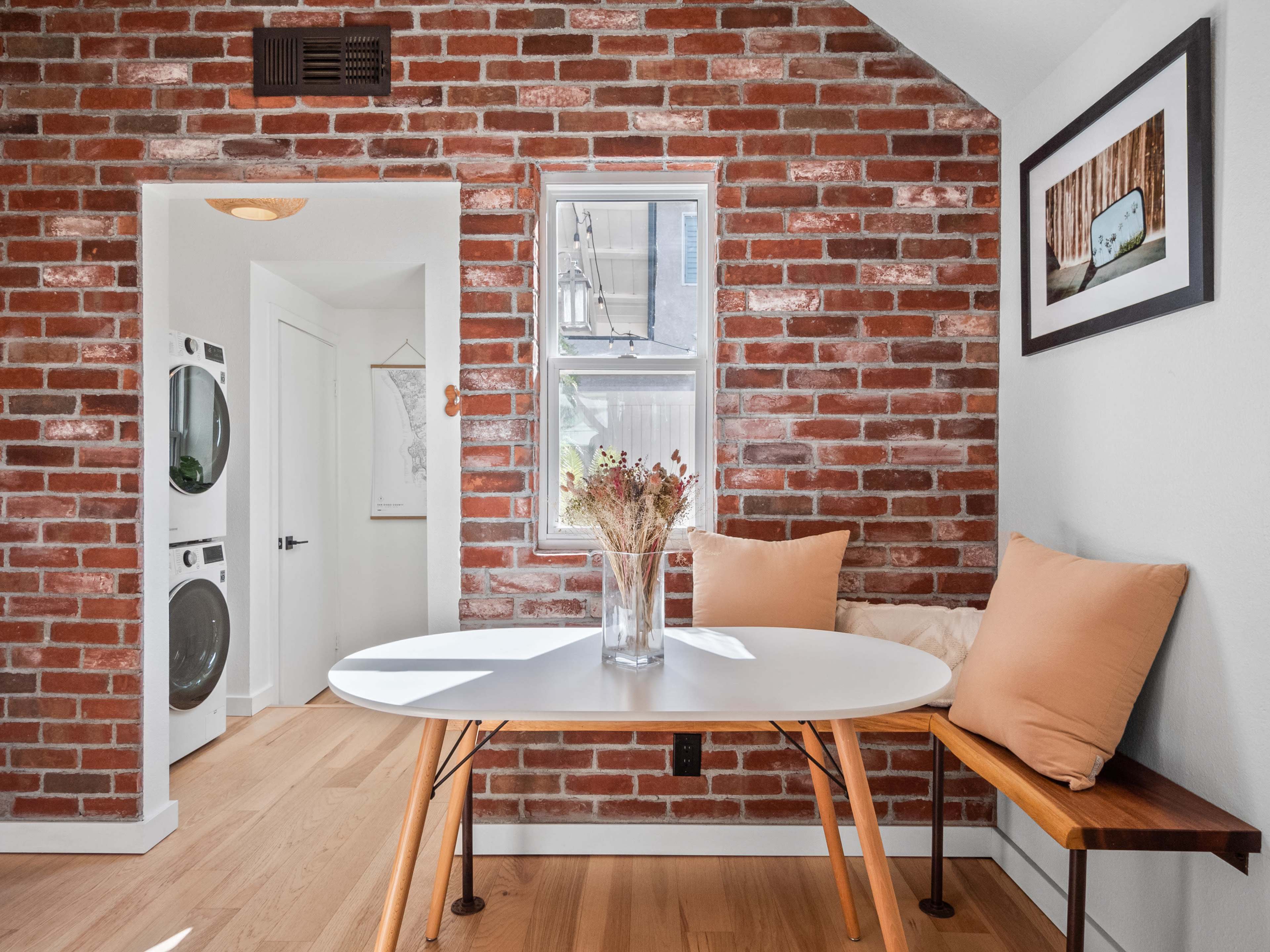 A small dining area features a round white table and a bench against a brick wall, with a vase of dried flowers and a framed picture on the wall.