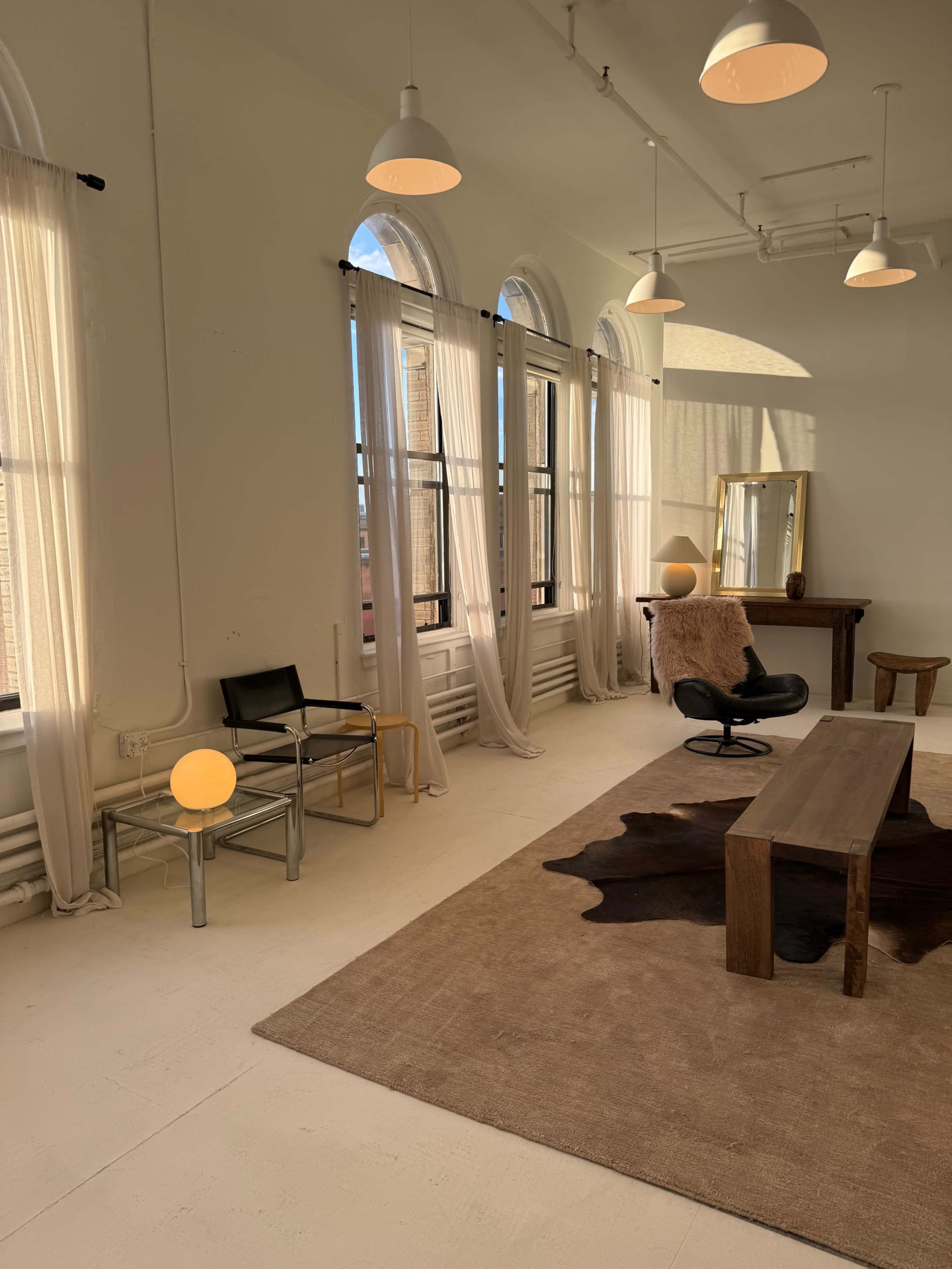 The image shows a bright, minimalist room with large windows, a wooden table, a chair, a mirror, and a light fixture.