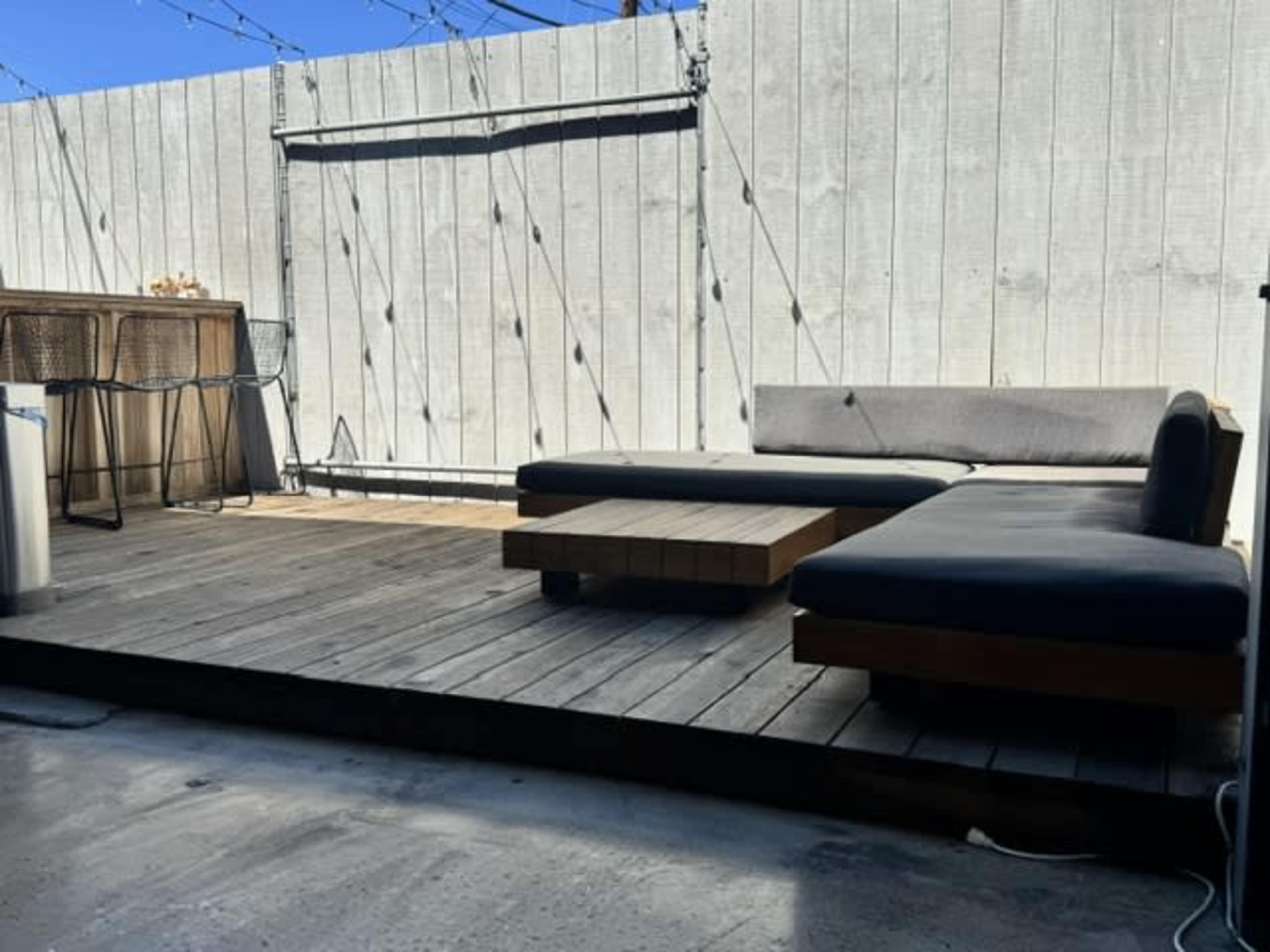 The image shows a wooden patio area with a low coffee table and a sectional couch against a gray wooden fence, with a bar area visible in the background.