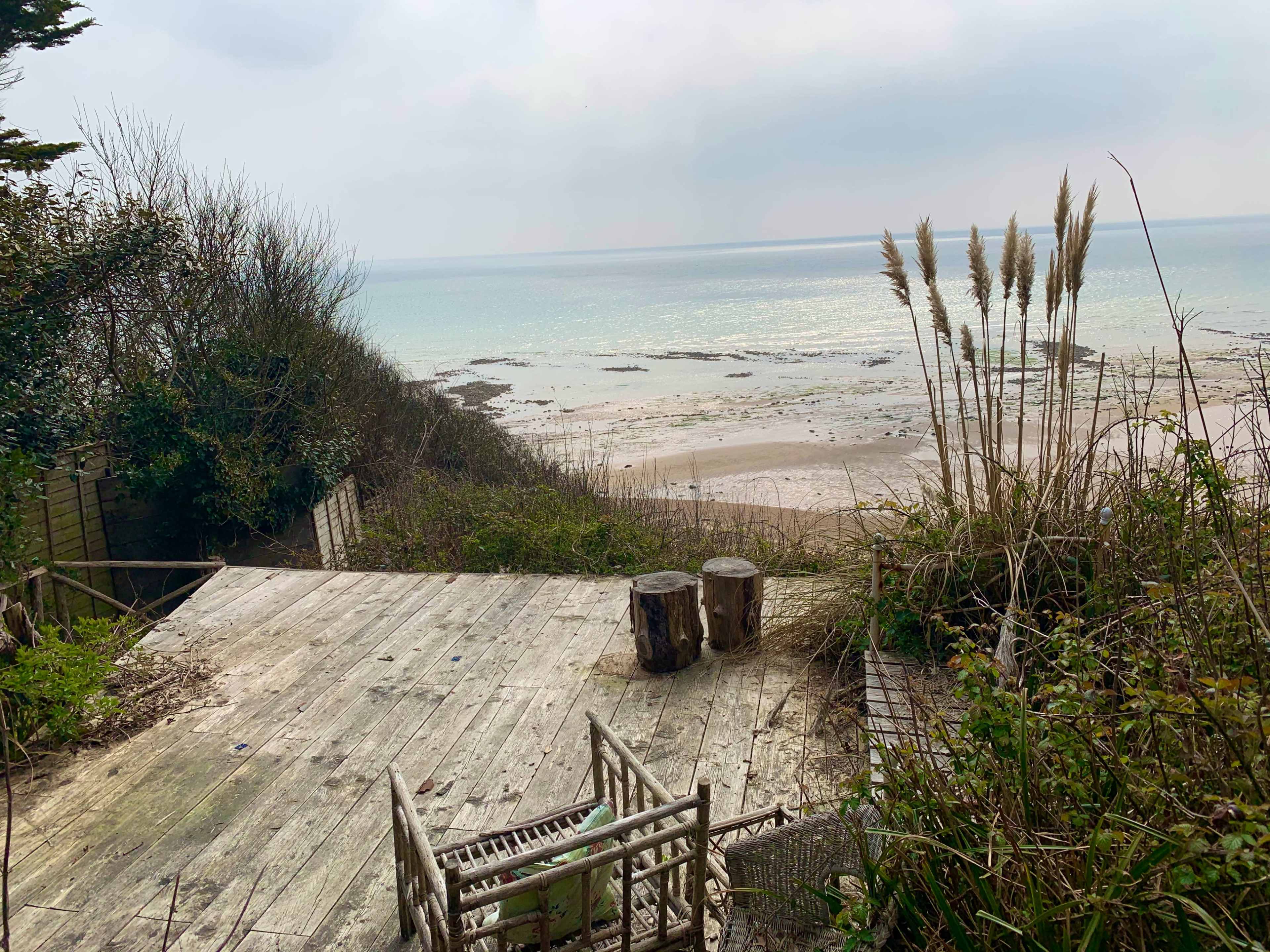 A weathered wooden deck overlooks a sandy beach and the ocean, surrounded by overgrown vegetation and coastal grass.