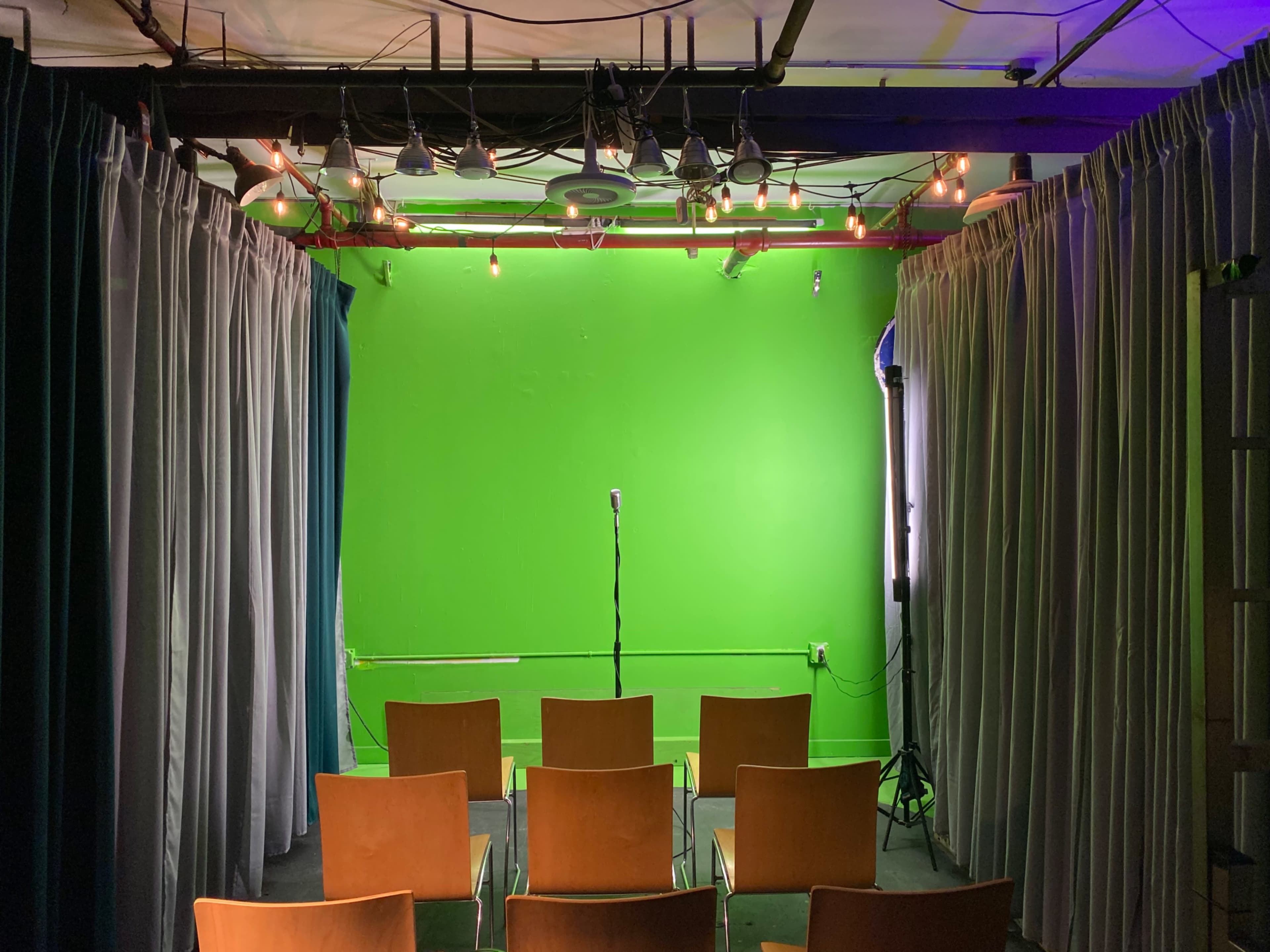 A small performance space with a green wall, several wooden chairs arranged in rows, and a microphone on a stand in the center.