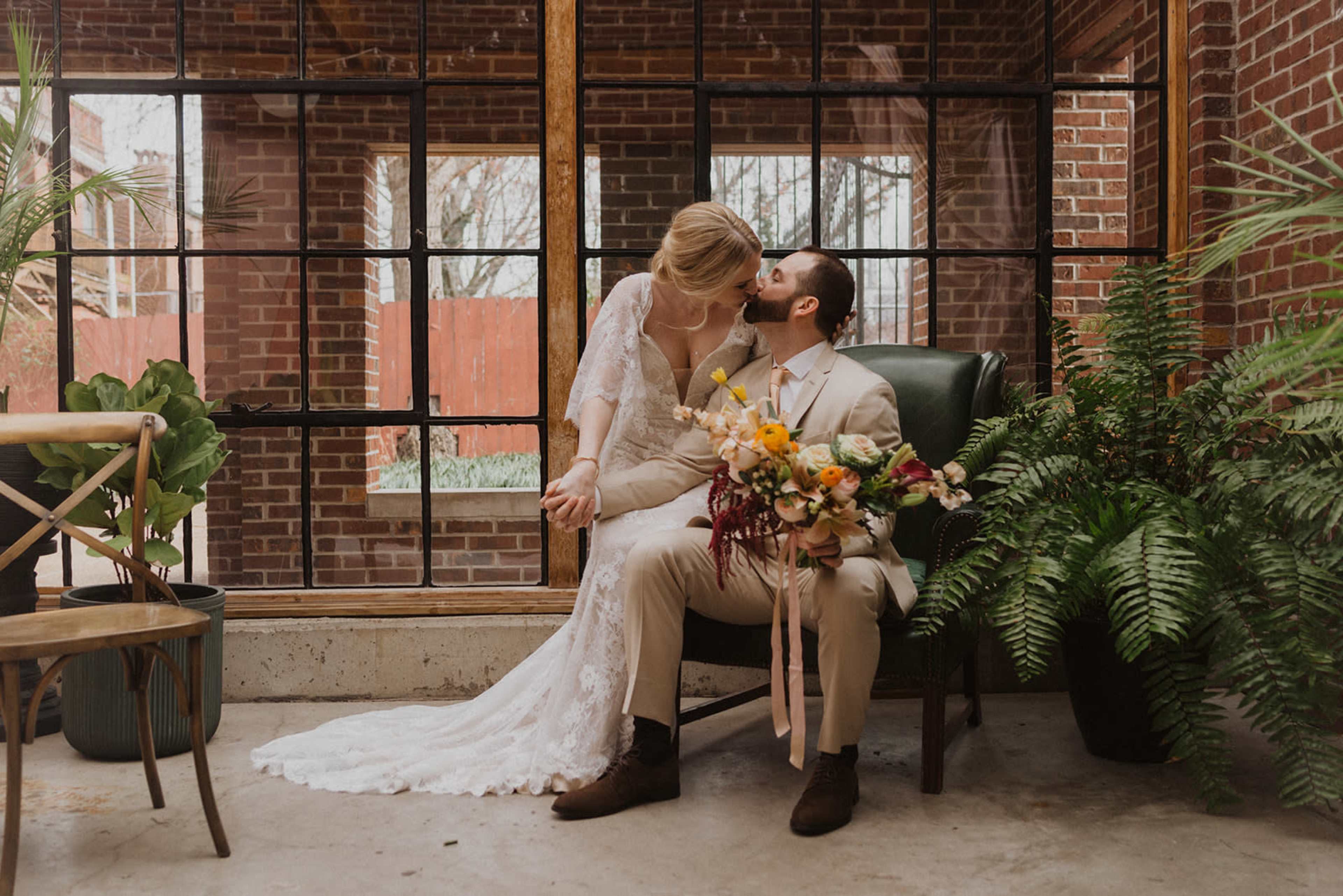 A bride and groom sit together in a plant-filled room, with the bride holding the groom's face as he holds a bouquet of flowers.