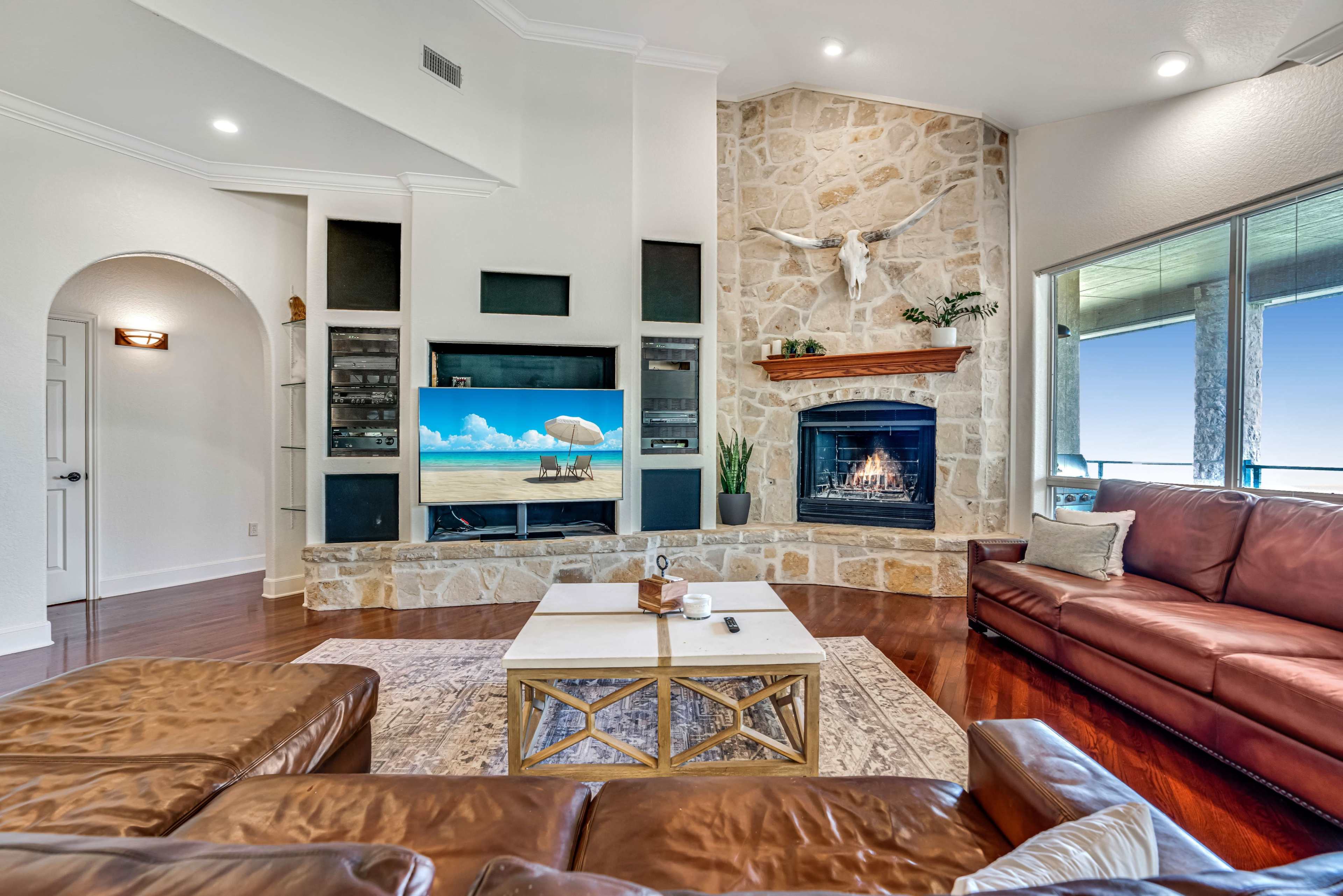 A modern living room with a stone fireplace, a large TV mounted on the wall, and a brown leather sofa arranged around a coffee table.