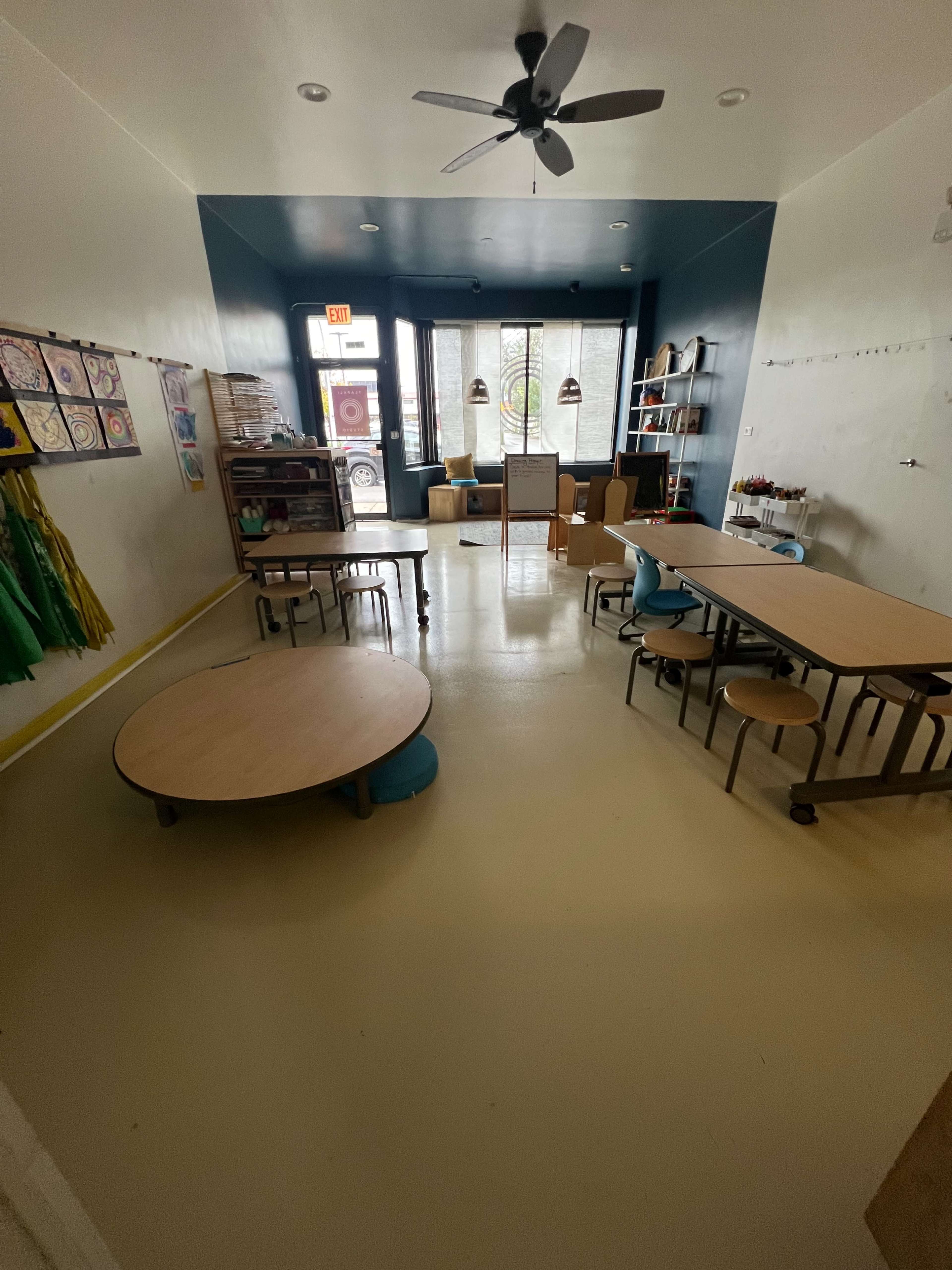 The image shows a well-lit classroom featuring various tables, chairs, and art supplies, with large windows allowing natural light to enter.