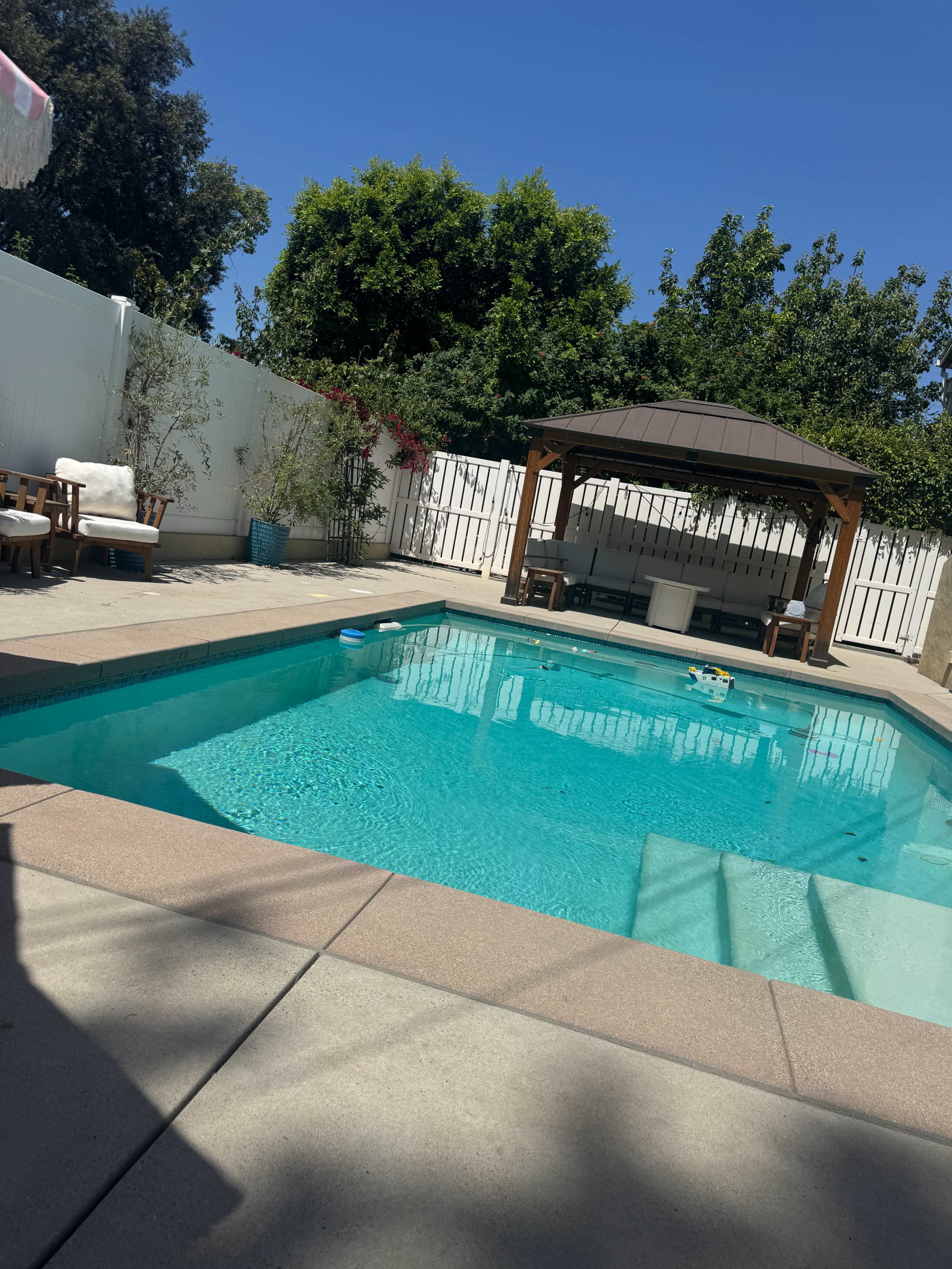 The image shows a backyard swimming pool with a gazebo, surrounded by a paved patio and lush greenery.