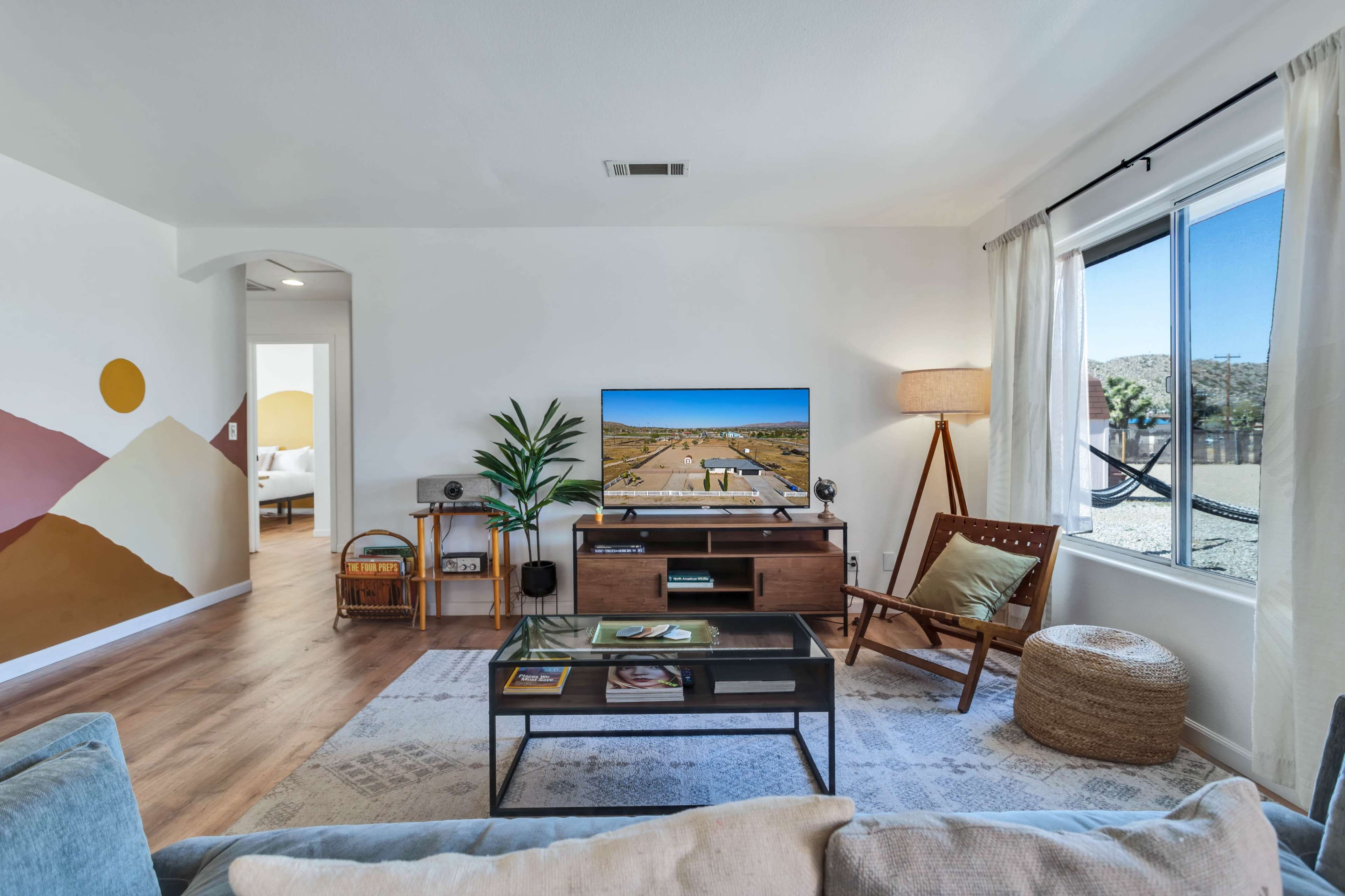 A modern living room features a television on a wooden stand, a plant by the window, and a chair beside a floor lamp, with a colorful mural on the wall and a view of the outdoors.