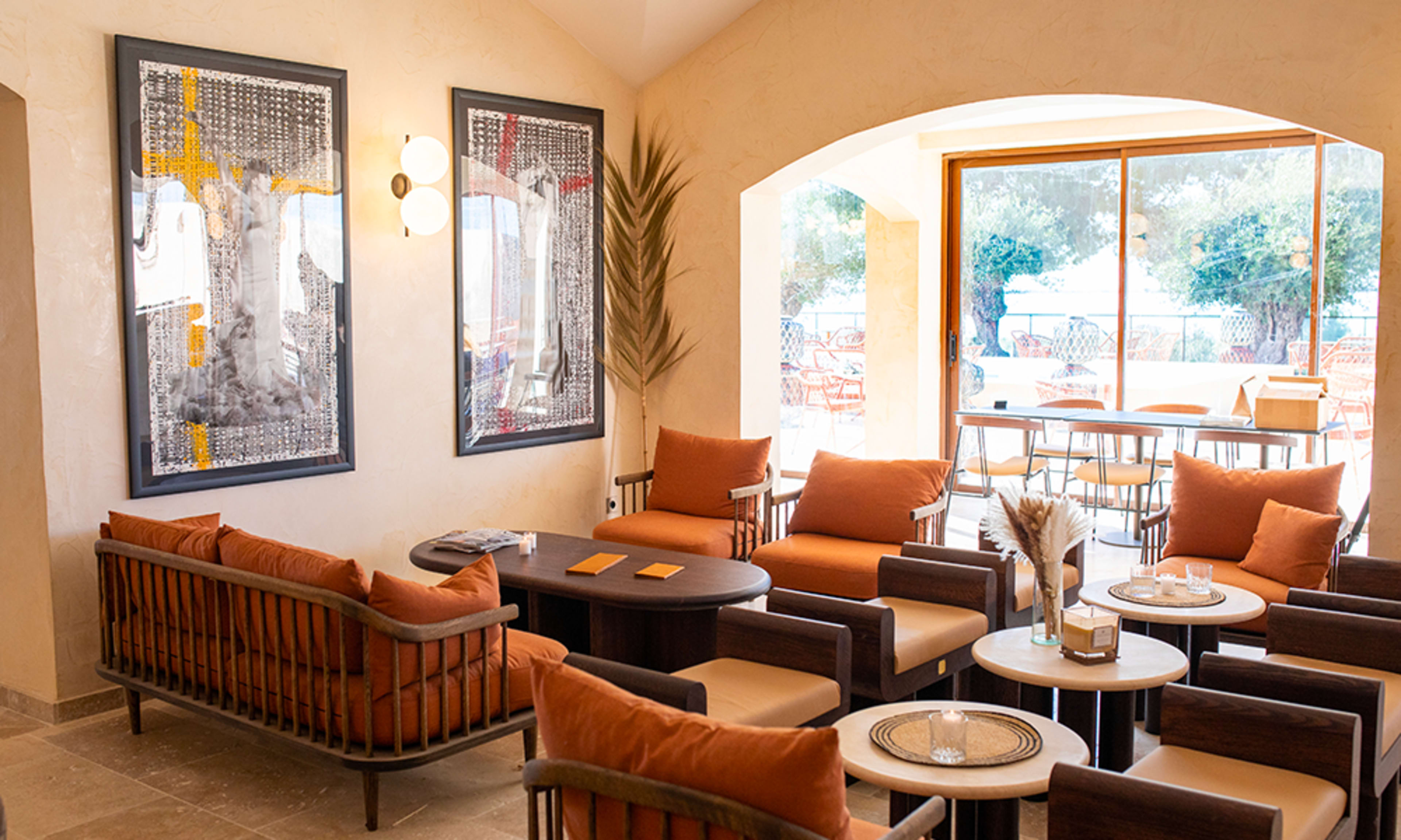 The image shows a modern lounge area with orange cushions on wooden sofas, two framed artworks on the wall, and large windows providing a view of an outdoor seating space.