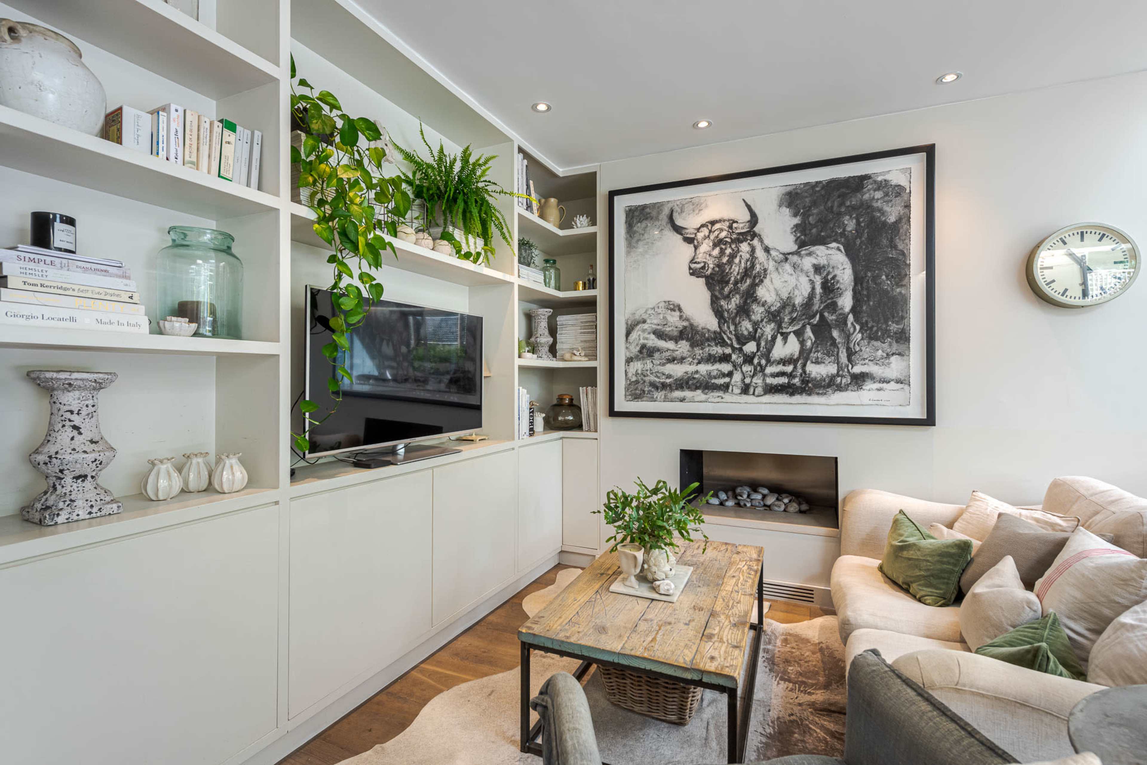A living room features a beige sofa, a wooden coffee table, and a large black-and-white bull artwork on the wall, surrounded by shelves with decorative items and plants.