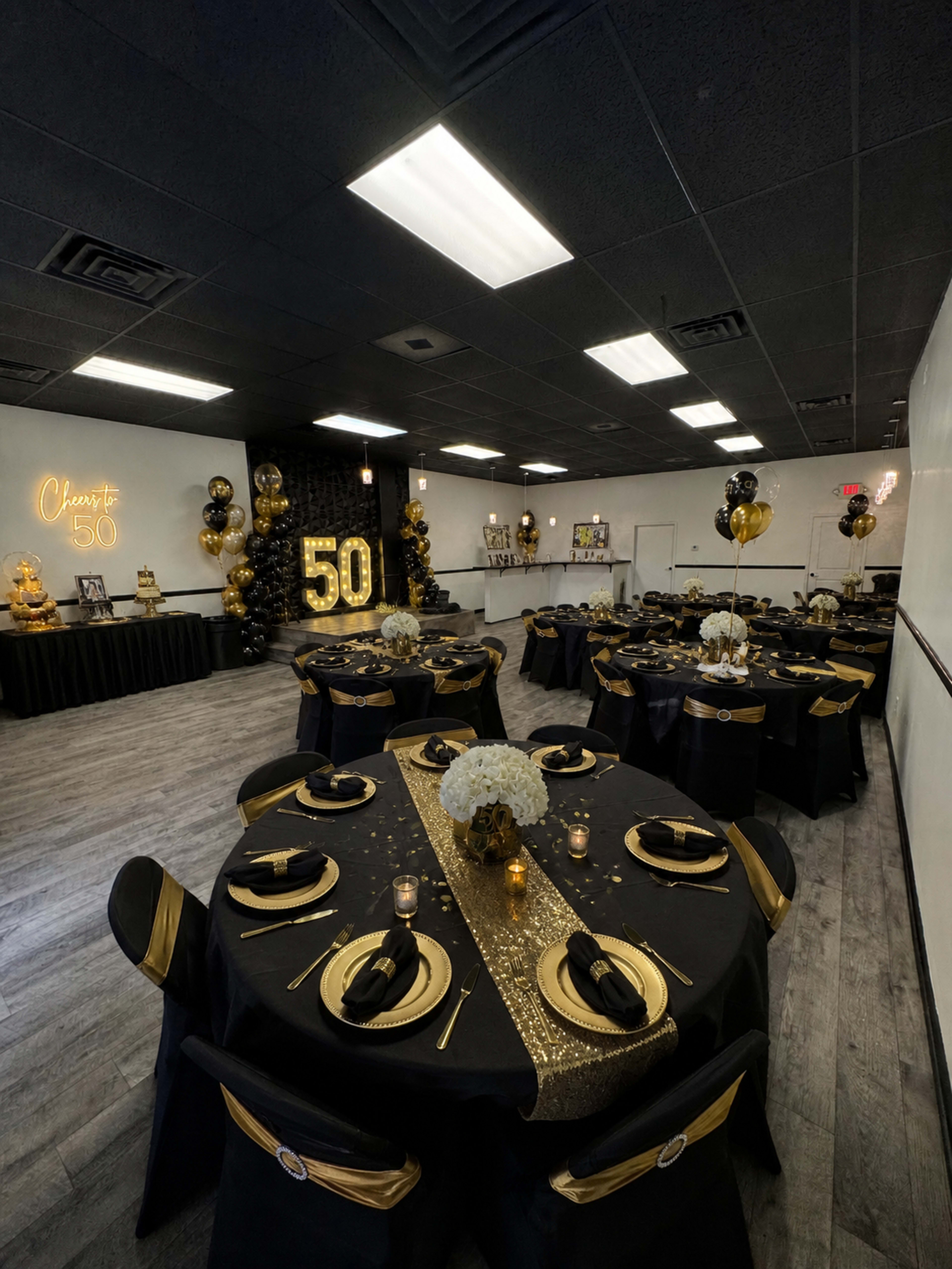 The image shows a banquet hall decorated for a 50th birthday celebration, featuring black and gold table settings, balloons, and a backdrop displaying the number "50."