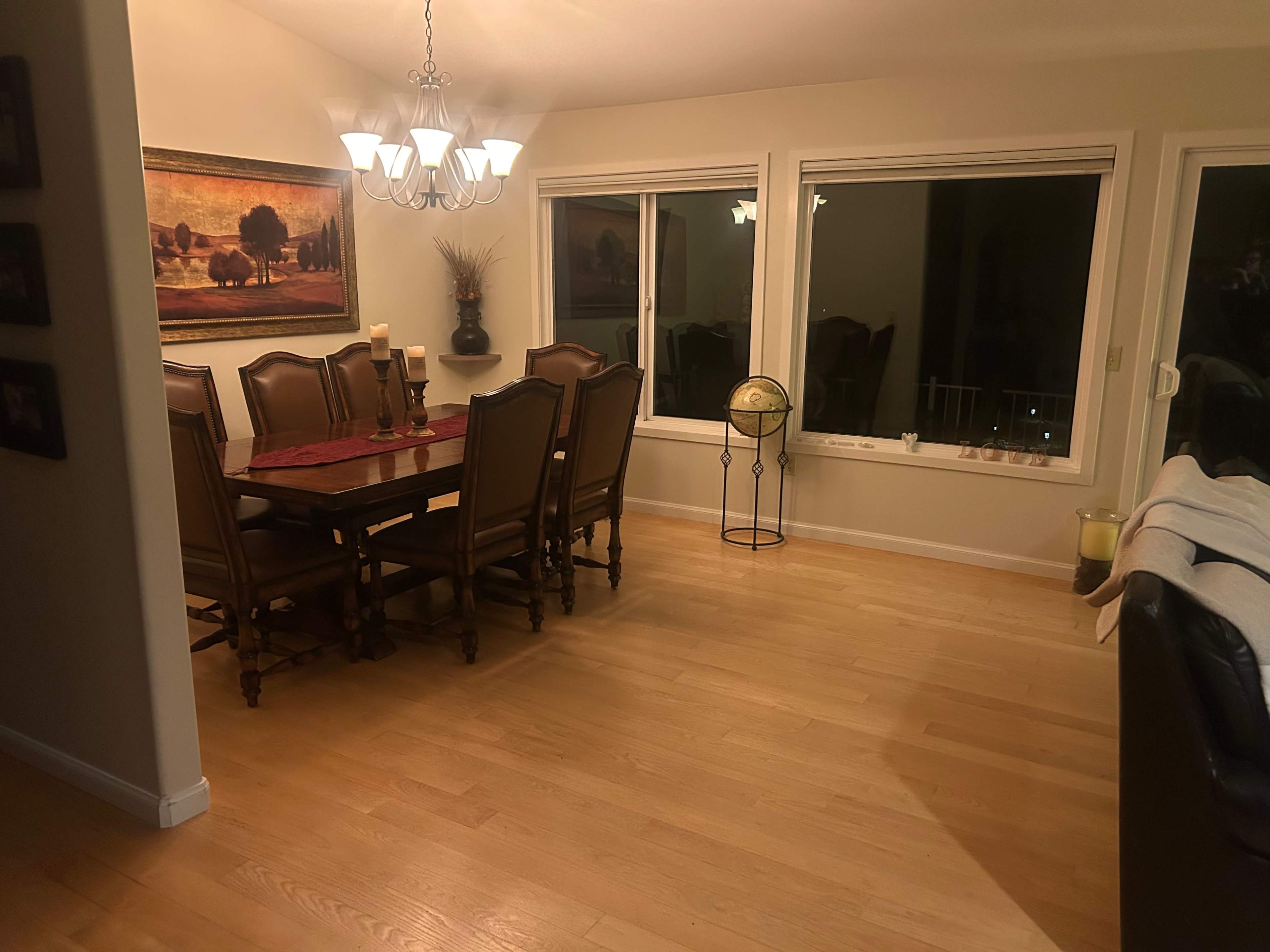 The image shows a dining room with a rectangular table surrounded by eight chairs, a chandelier above the table, and large windows allowing light into the space.