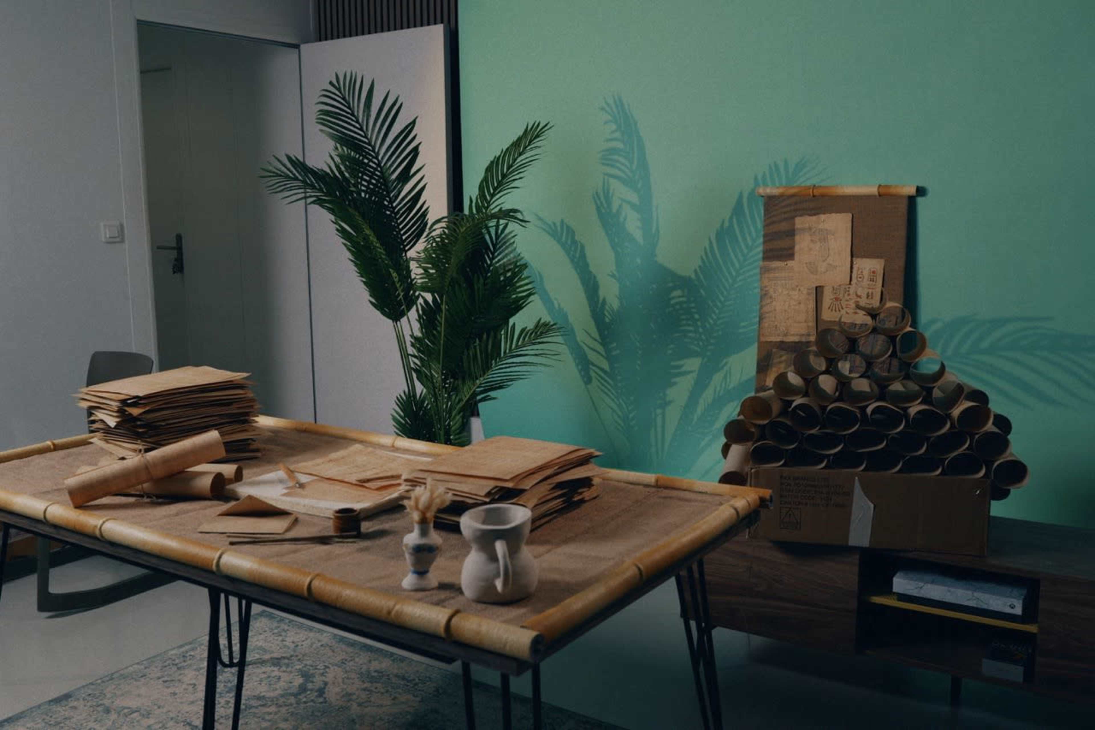 The image shows a room with a table covered in sheets of paper and a plant, alongside a collection of cardboard tubes stacked in a box against a green wall.