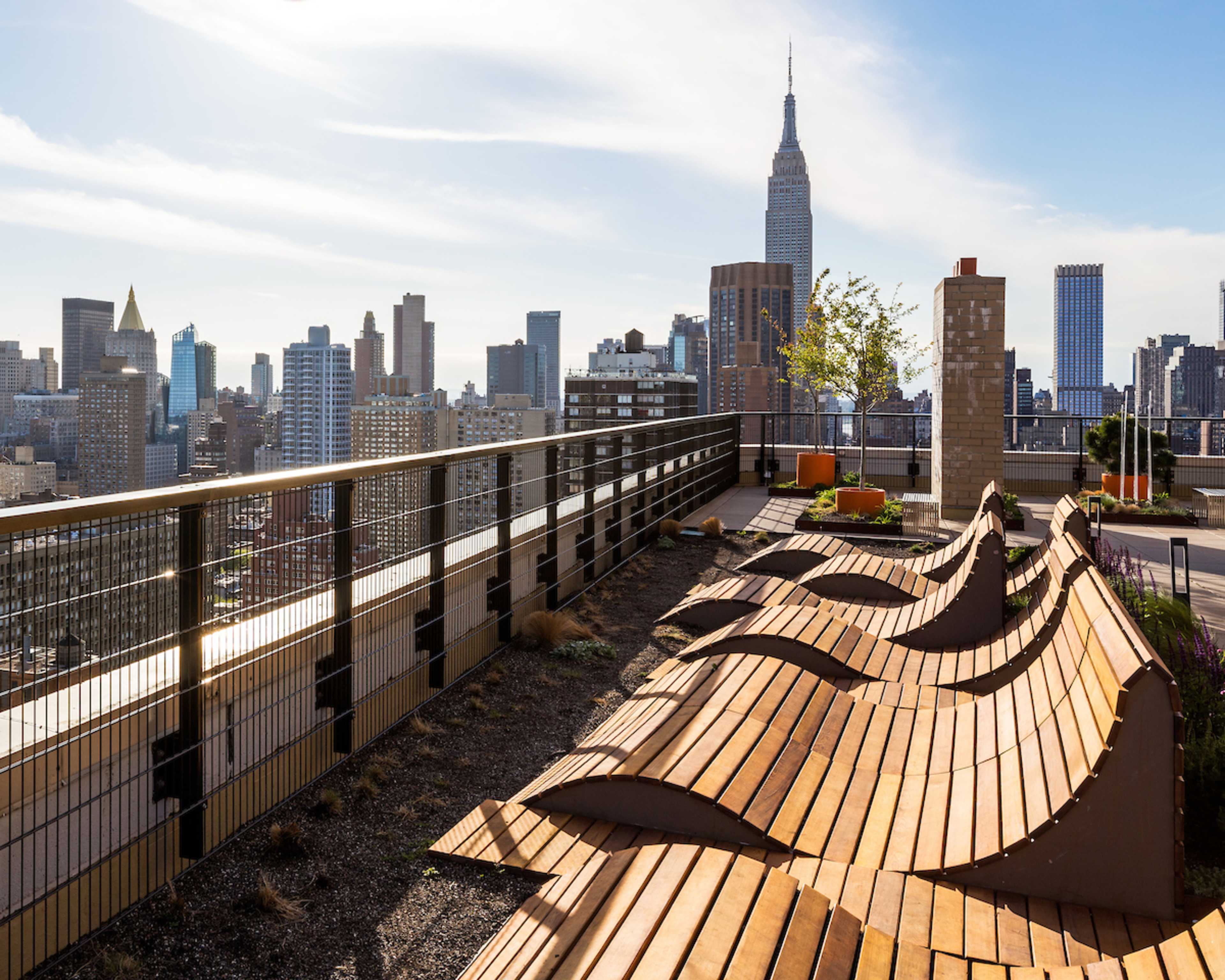 The image shows a rooftop terrace with wooden seating and a view of the Manhattan skyline, including the Empire State Building.