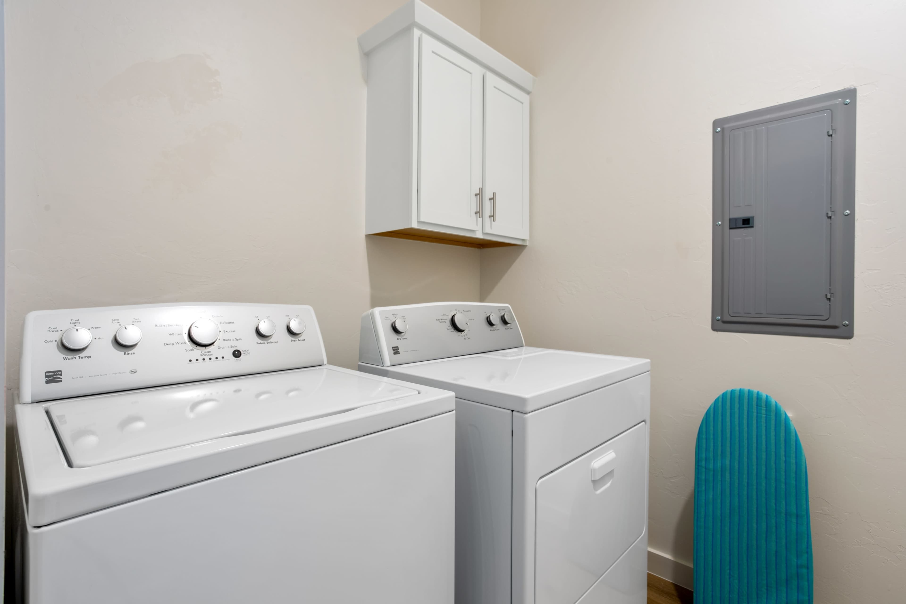 A laundry room with a washing machine, dryer, a wall-mounted electrical panel, and a light blue ironing board.