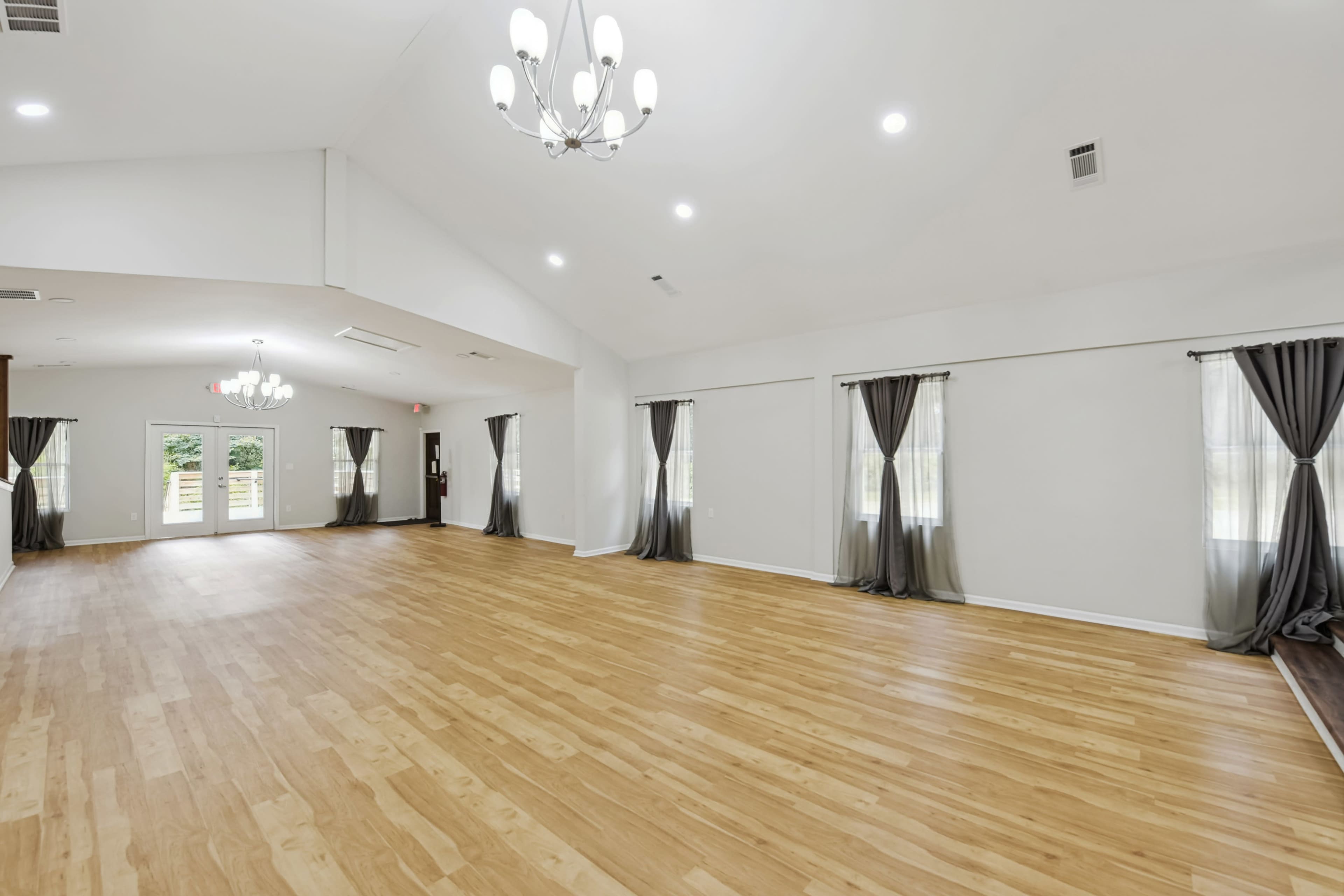 The image shows a spacious, brightly lit room with wooden flooring, large windows covered by curtains, and a high ceiling with chandeliers.