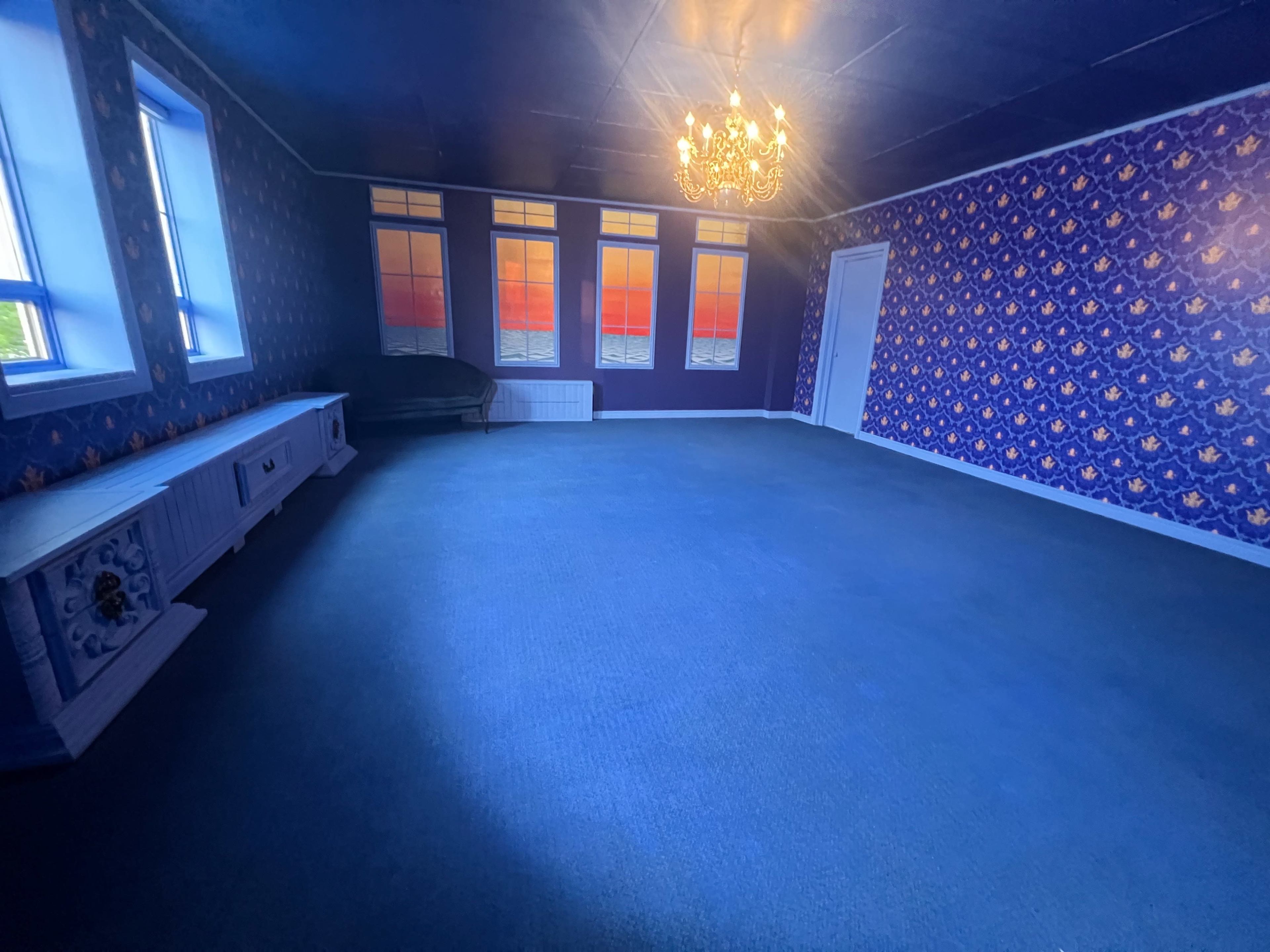 The room features blue walls with floral wallpaper, a chandelier, large windows with sunset views, and a carpeted floor.