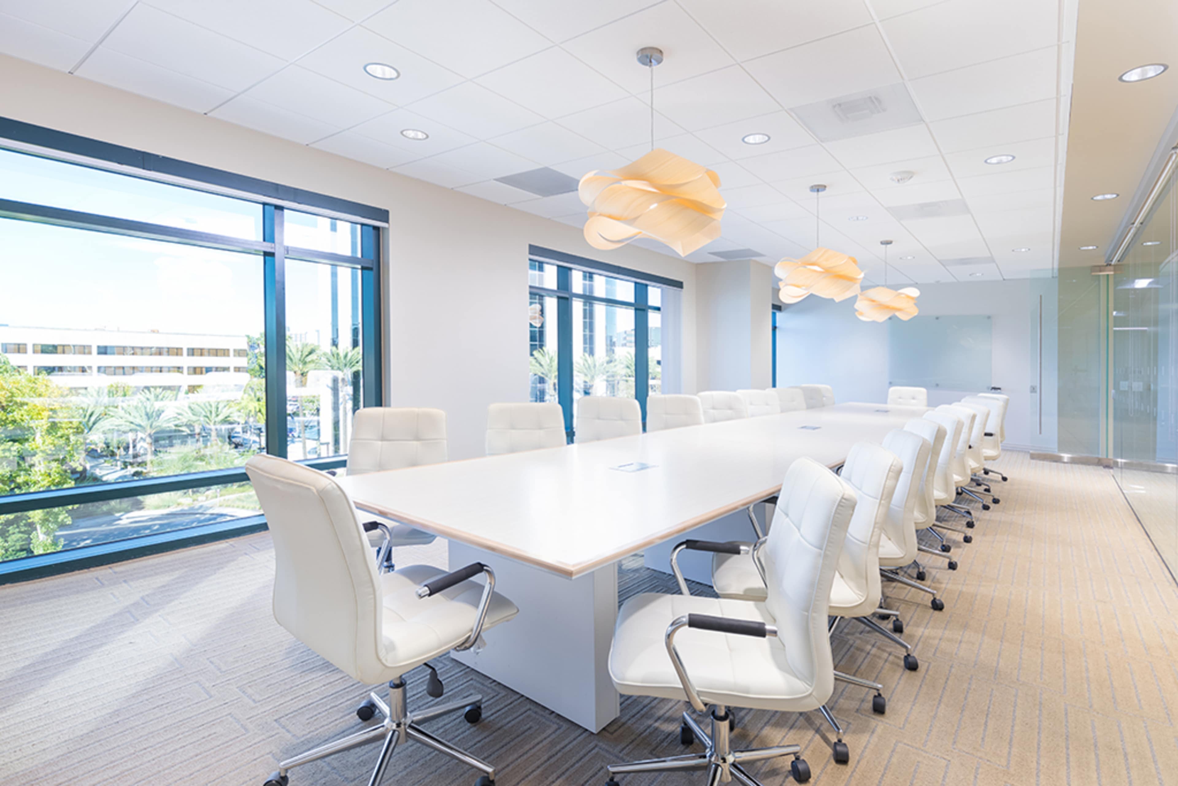 A modern conference room with a long white table surrounded by ergonomic white chairs, illuminated by pendant lights and featuring large windows overlooking an outdoor landscape.