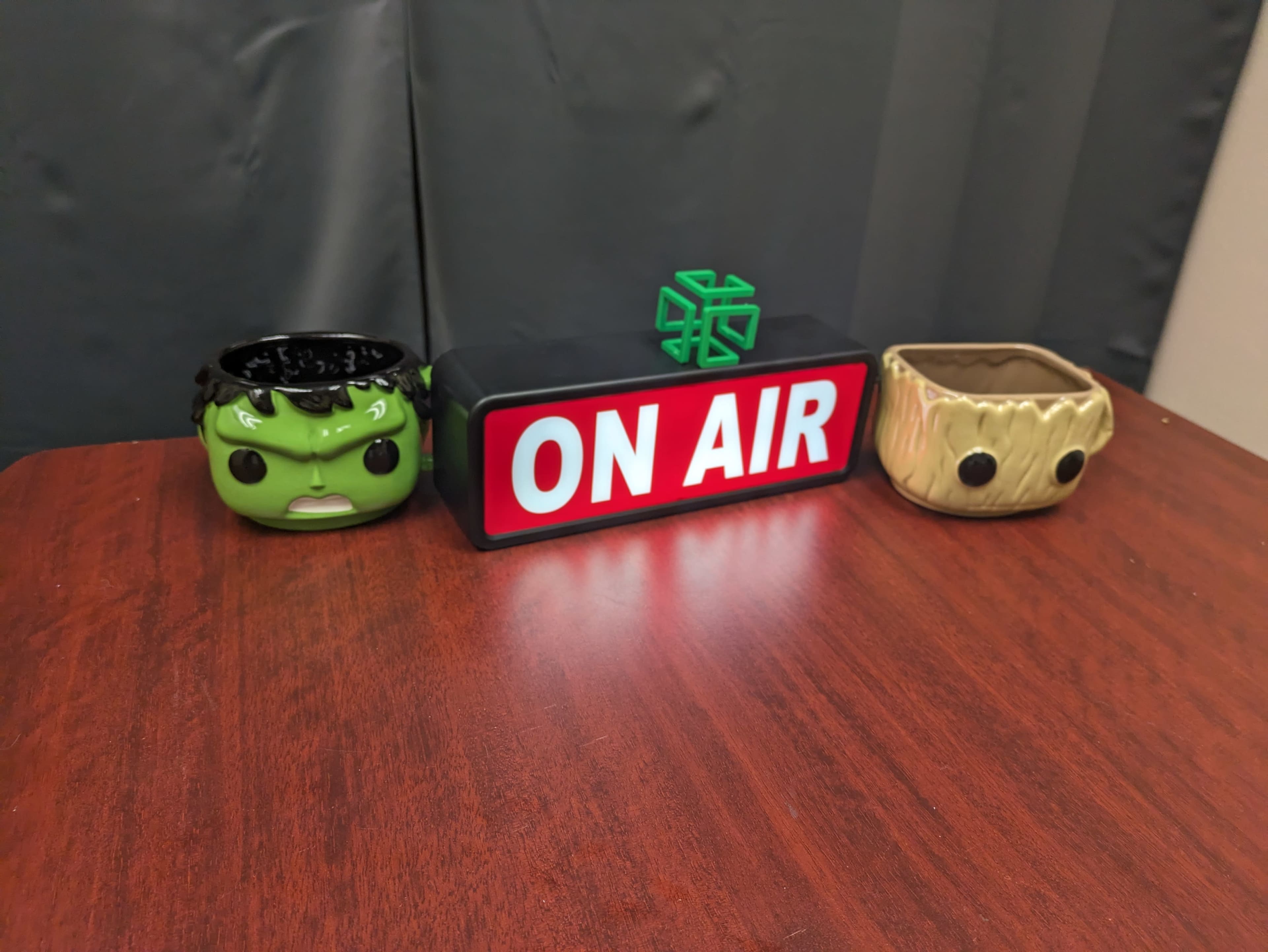 The image shows a wooden table with a red and white "ON AIR" sign centered between two decorative planters styled as comic characters.