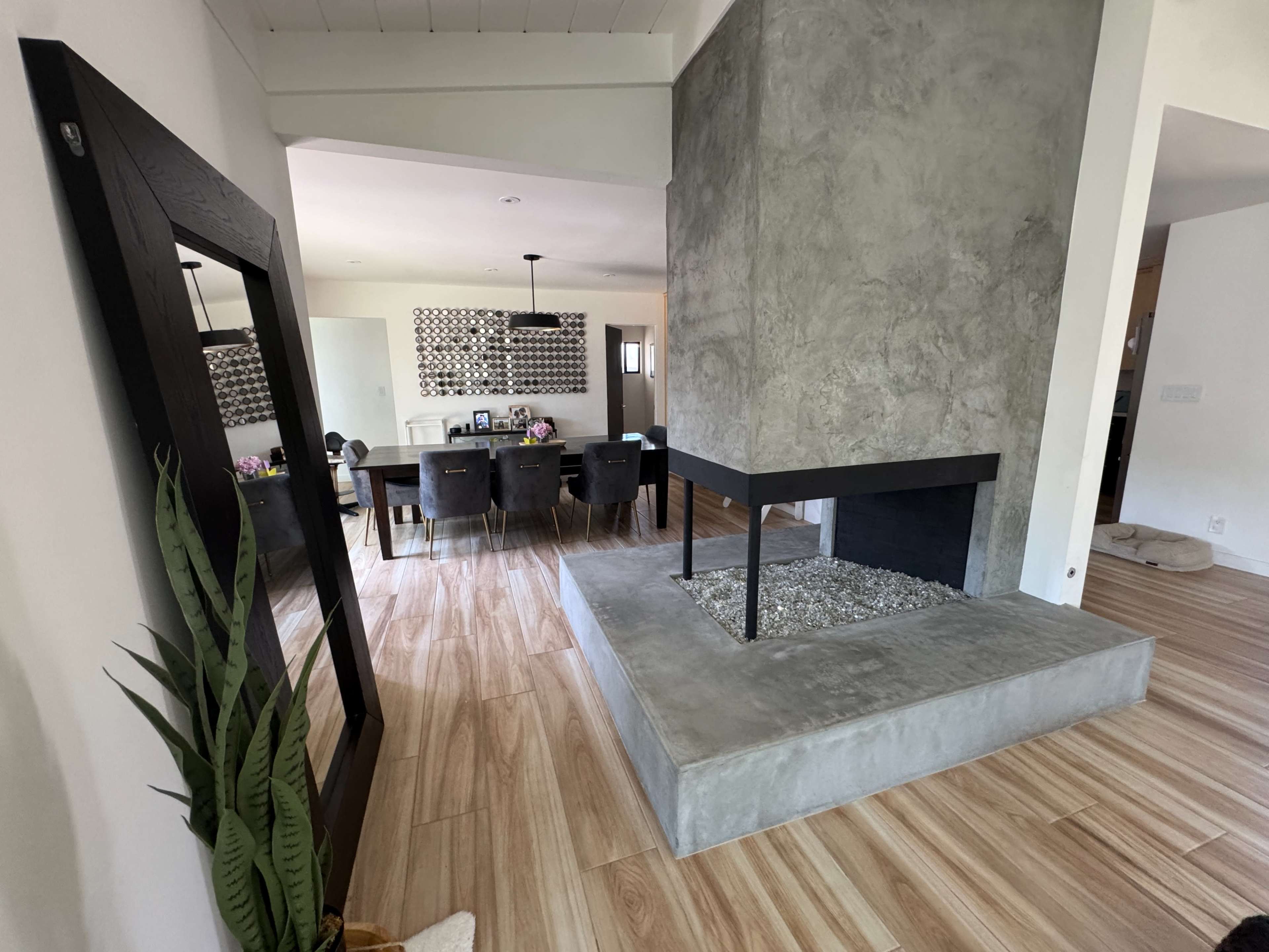 The image shows a modern living space featuring a concrete fireplace, a large mirror, and a dining area in the background with a table and chairs.