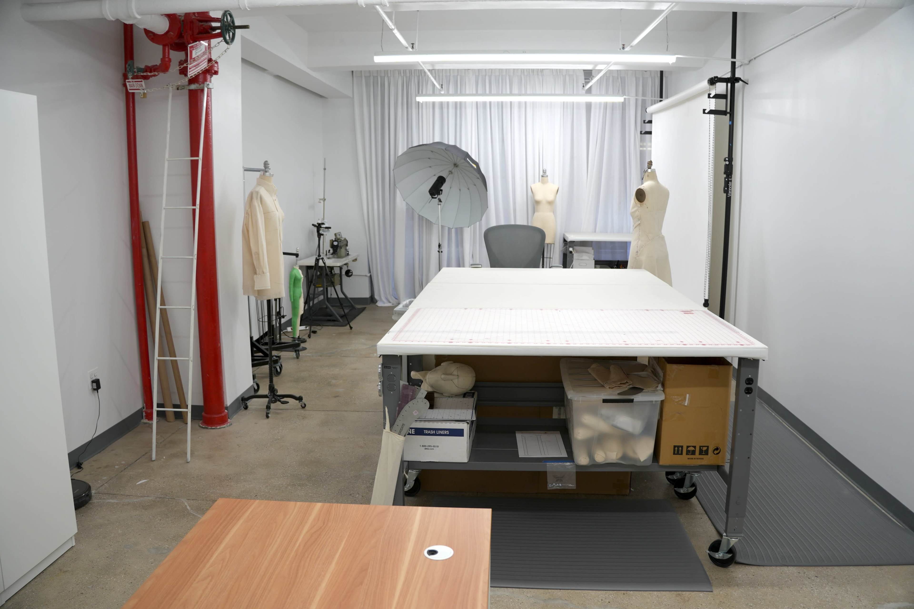 The image shows a spacious design studio featuring a large cutting table, fashion mannequins, and various sewing tools organized around the area.